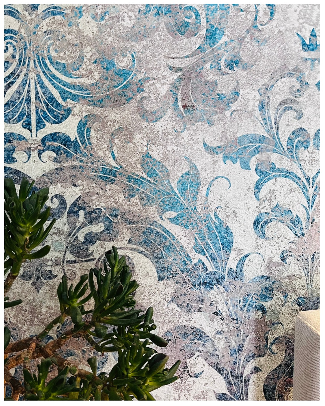 Dekoracyjna ściana z wzorem roślinnym w odcieniach błękitu i srebra, z fragmentem zielonej rośliny w lewym dolnym rogu. Efektowna tapeta z fakturą.
