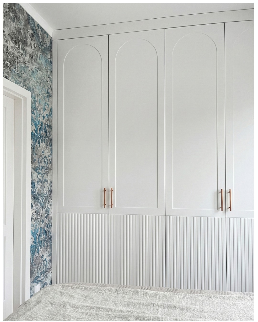 Biała szafa wnękowa z frezowanymi frontami i łukowatymi górnymi krawędziami, uzupełniona wzorzystą tapetą w odcieniach szarości i błękitu, widok z dołu.
