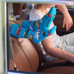 HOLA STUDIO SPÓŁKA Z OGRANICZONĄ ODPOWIEDZIALNOŚCIĄ - Nogi w błękitnych skarpetkach z motywem samochodów PRL-u, wystawione przez okno starego auta w Chludowie, brązowa skórzana tapicerka siedzenia