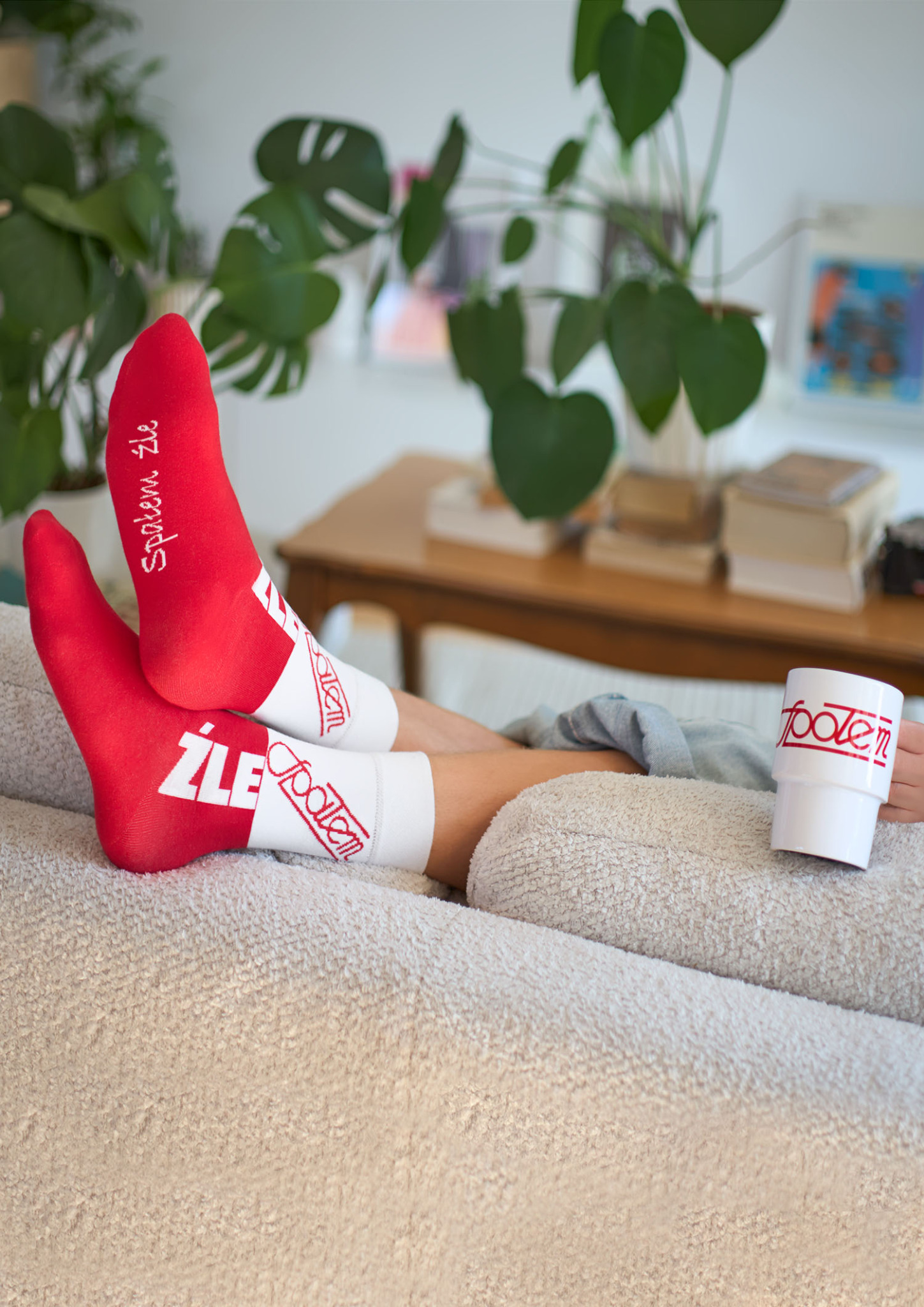 Osoba relaksuje się na kanapie w czerwono-białych skarpetkach z napisem oraz trzyma kubek z logo, w tle widoczna roślina Monstera i stolik.
