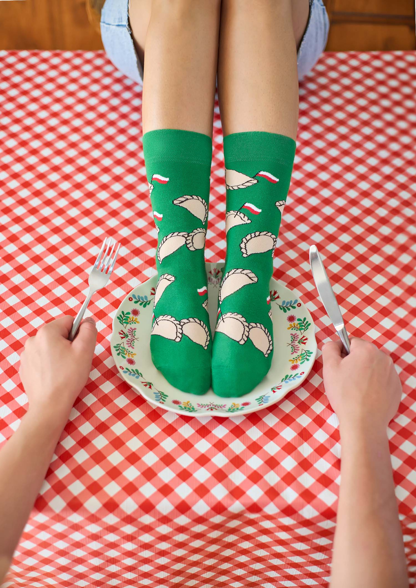 Nogi w zielonych skarpetkach z motywem pierogów i flagi Polski stojące na talerzu, dwie ręce trzymają nóż i widelec nad czerwono-białym obrusem w kratę.