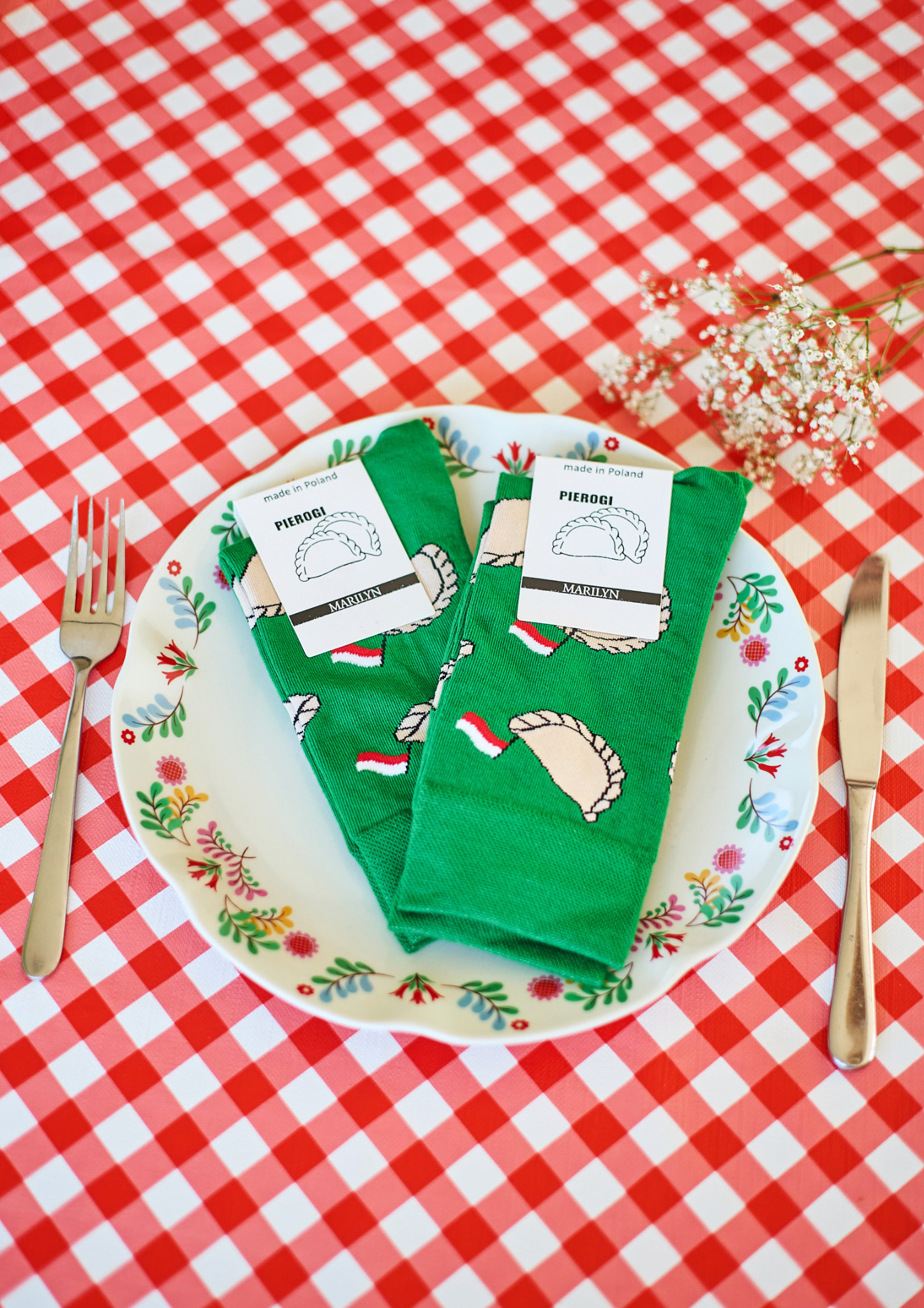 Zielone skarpetki z motywem pierogów leżą na talerzu z kwiatowym wzorem, obok sztućce i gałązka gipsówki na czerwono-białym obrusie w kratkę.