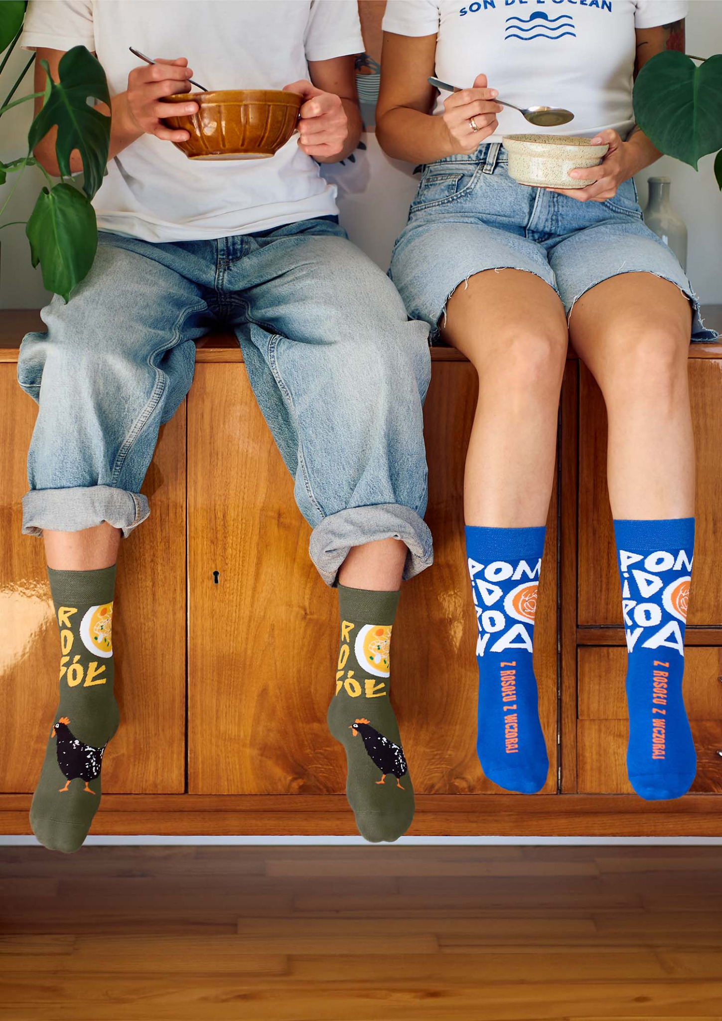 Dwie osoby siedzą na drewnianym kredensie, jedząc z ceramicznych misek, eksponując zabawne skarpetki z motywem rosołu i kurczaka.