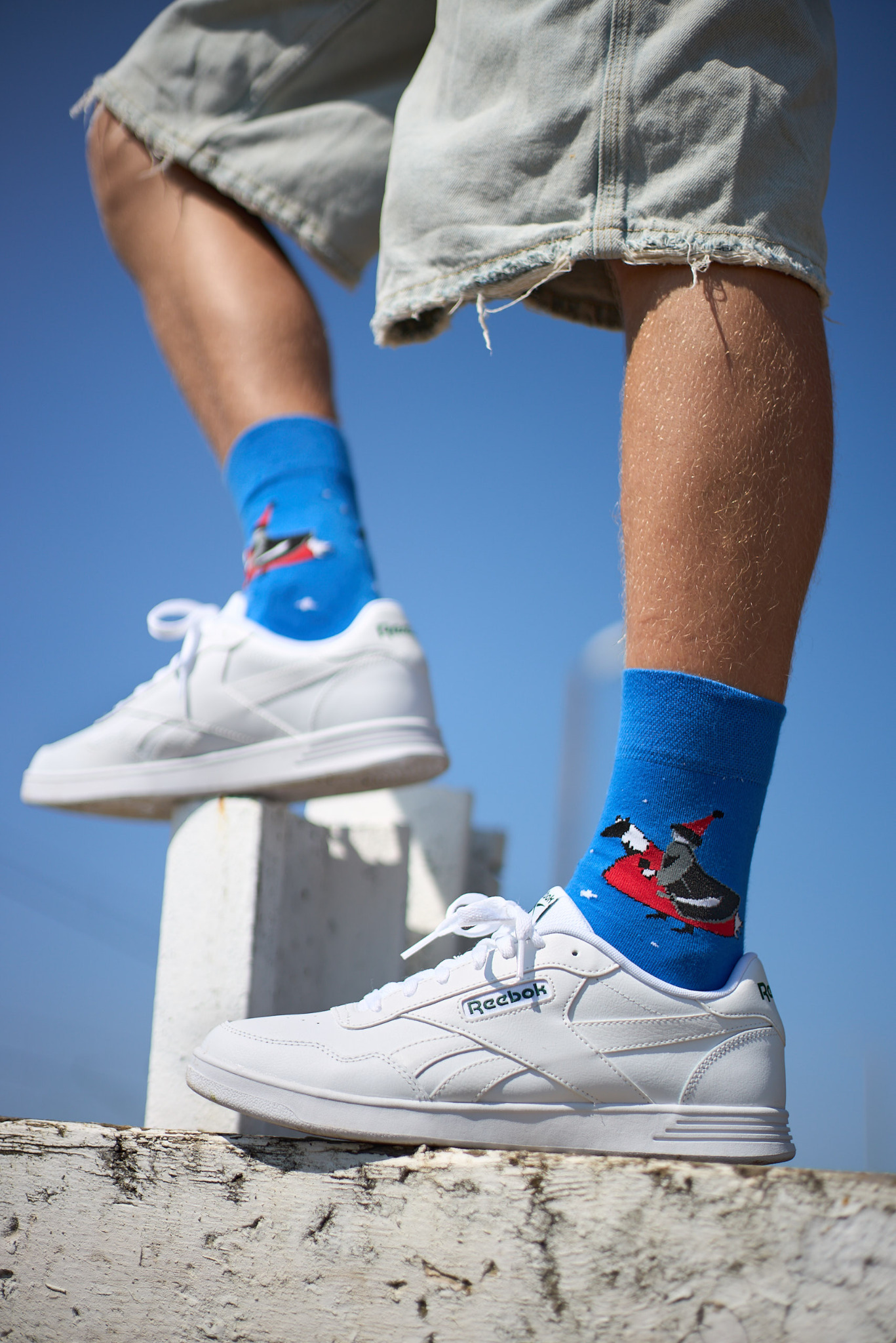 Męskie nogi w krótkich dżinsowych spodenkach i białych sneakersach Reebok, eksponujące niebieskie skarpetki z motywem pingwina na czerwonych sankach, na tle błękitnego nieba.