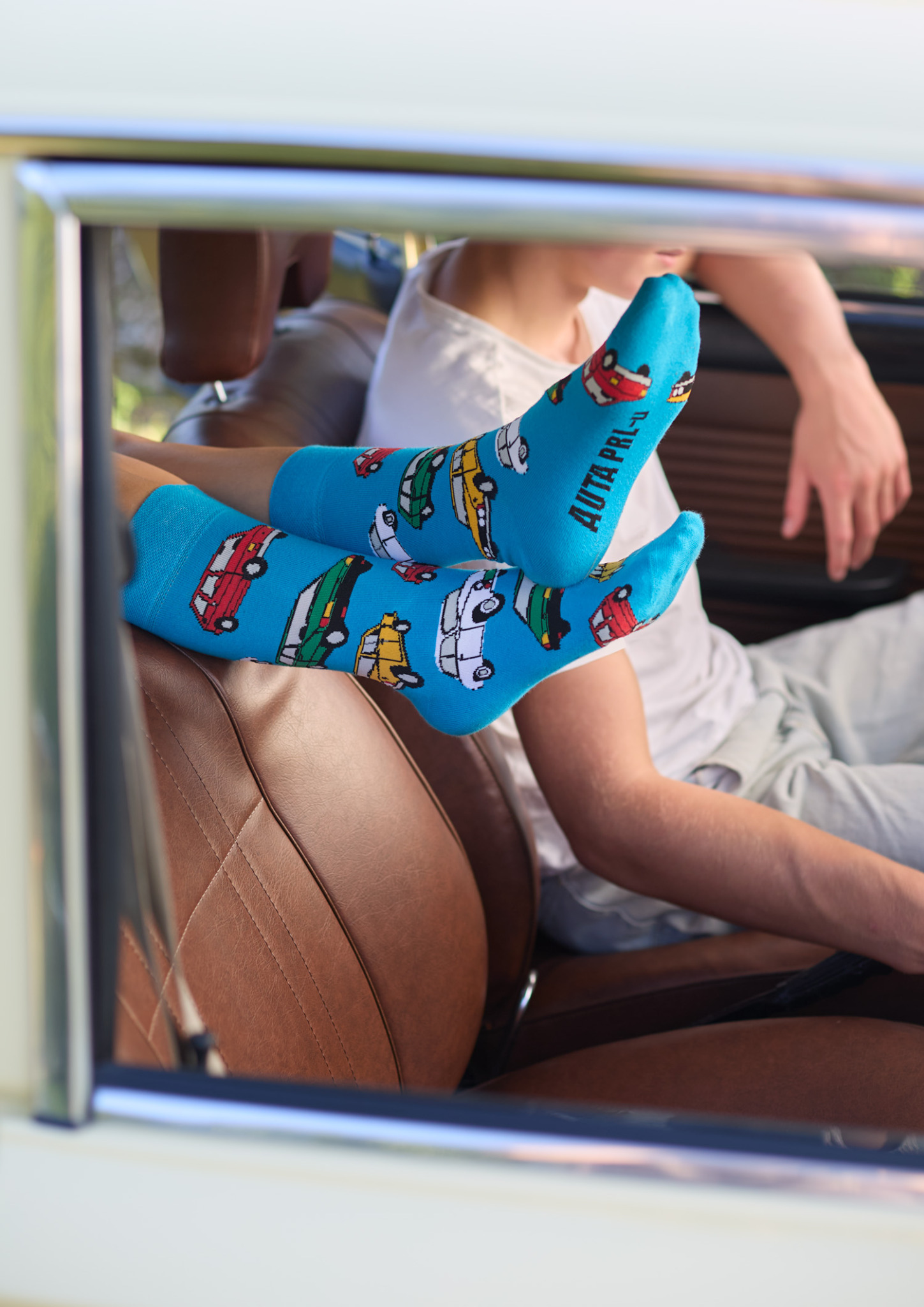 Nogi w błękitnych skarpetkach z motywem samochodów PRL-u, wystawione przez okno starego auta w Chludowie, brązowa skórzana tapicerka siedzenia