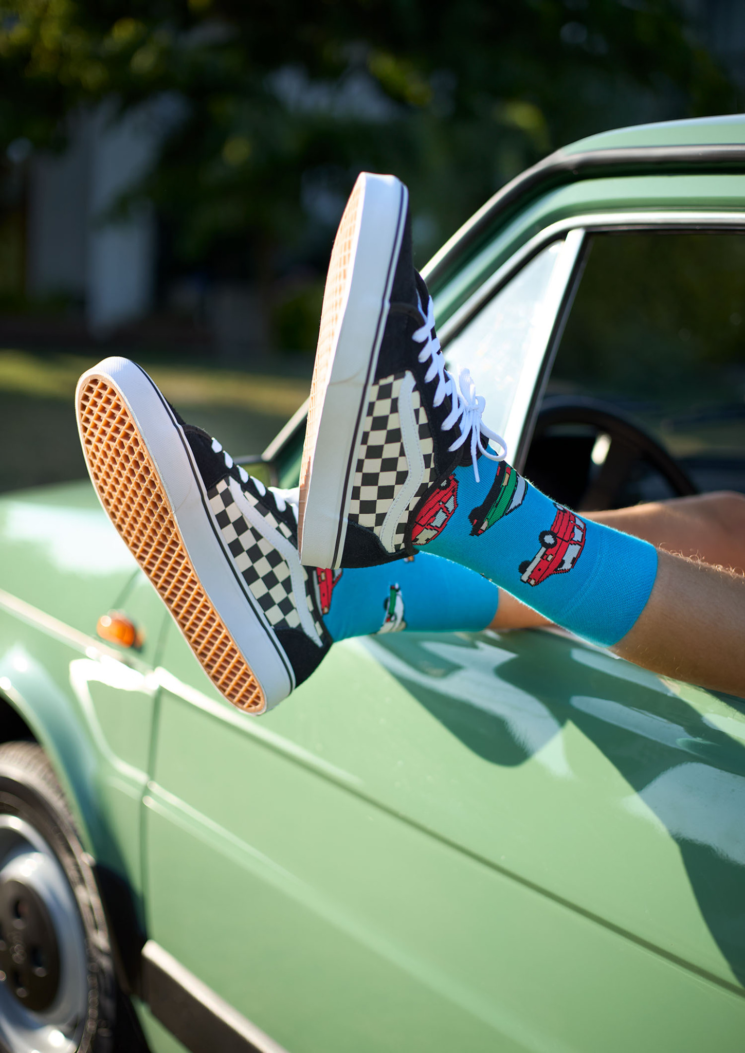 Osoba wystawia nogi w szachownicowych Vansach i niebieskich skarpetkach w samochodziki przez okno pistacjowego klasycznego auta.