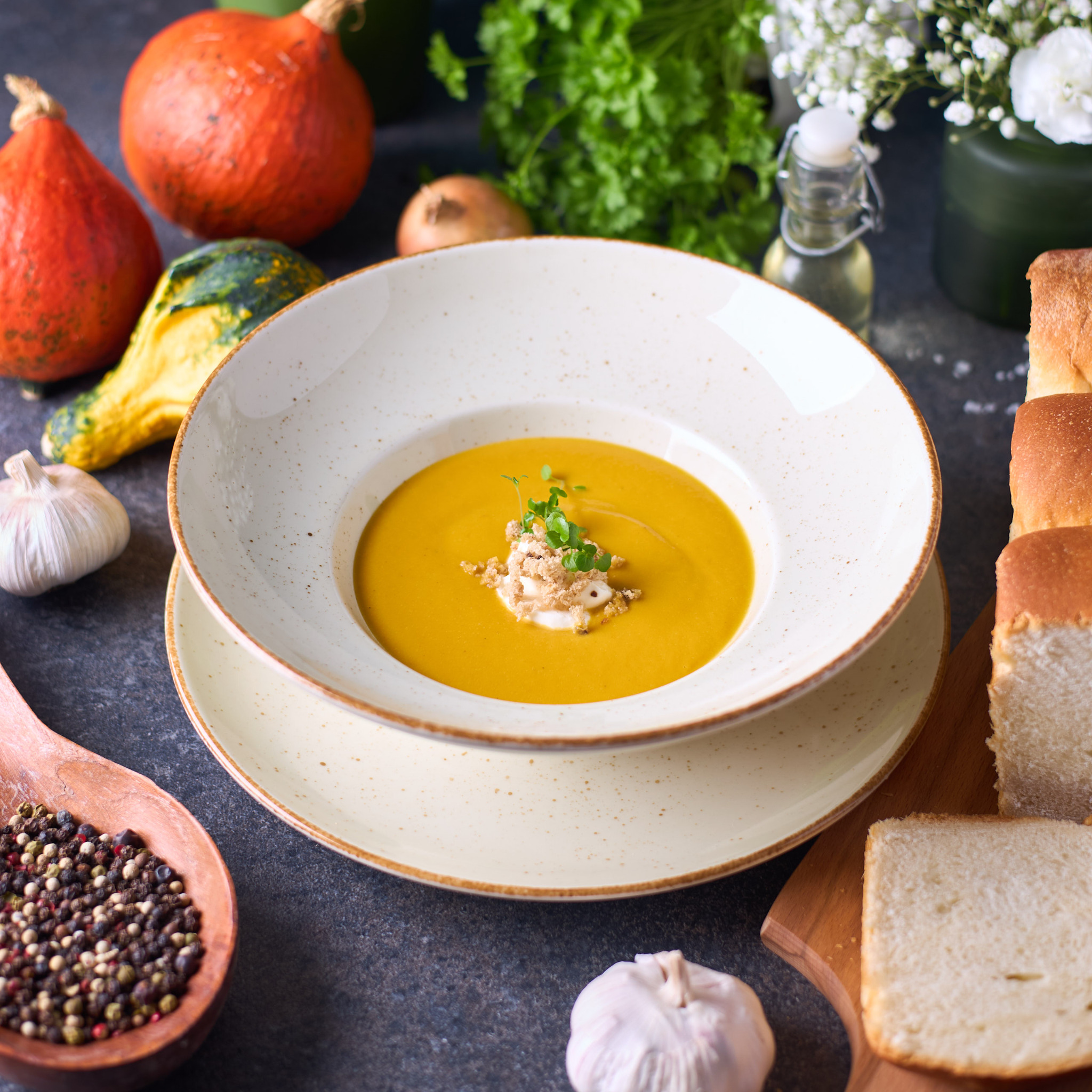 Krem z dyni w białym talerzu, udekorowany kiełkami i posypką, na tle dyni hokkaido, czosnku, pieprzu w drewnianej łyżce oraz pieczywa.