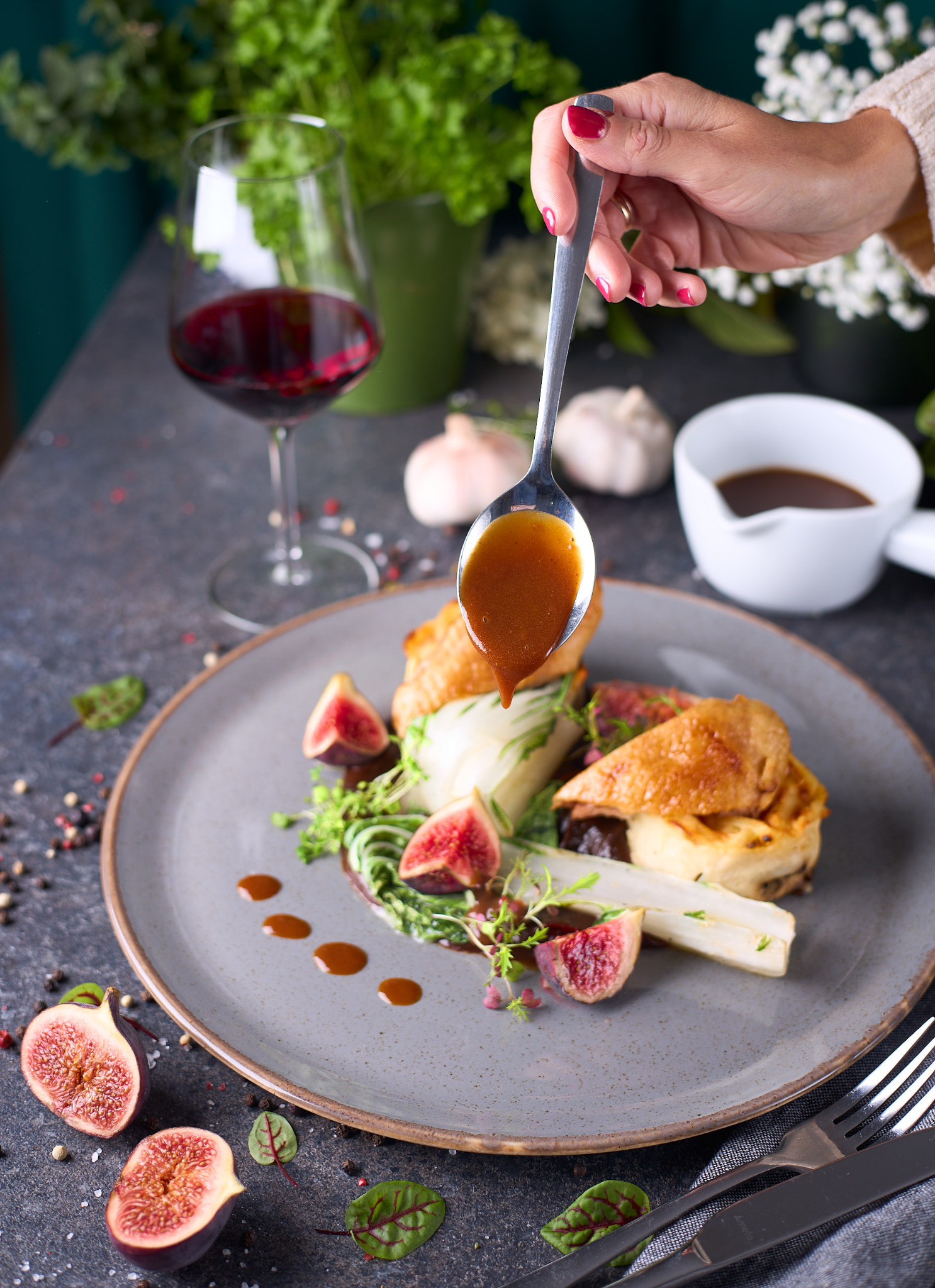 Elegancko podane danie z figami, sosem i białymi szparagami na szarym talerzu, polewane sosem z łyżki, w tle kieliszek czerwonego wina i czosnek.