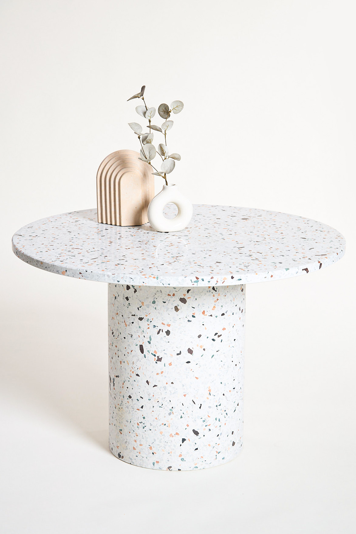 Okrągły stół z blatem i nogą w stylu lastryko, na nim dekoracyjny łuk z piaskowca i biały wazonik z gałązką eukaliptusa, na jasnym tle.