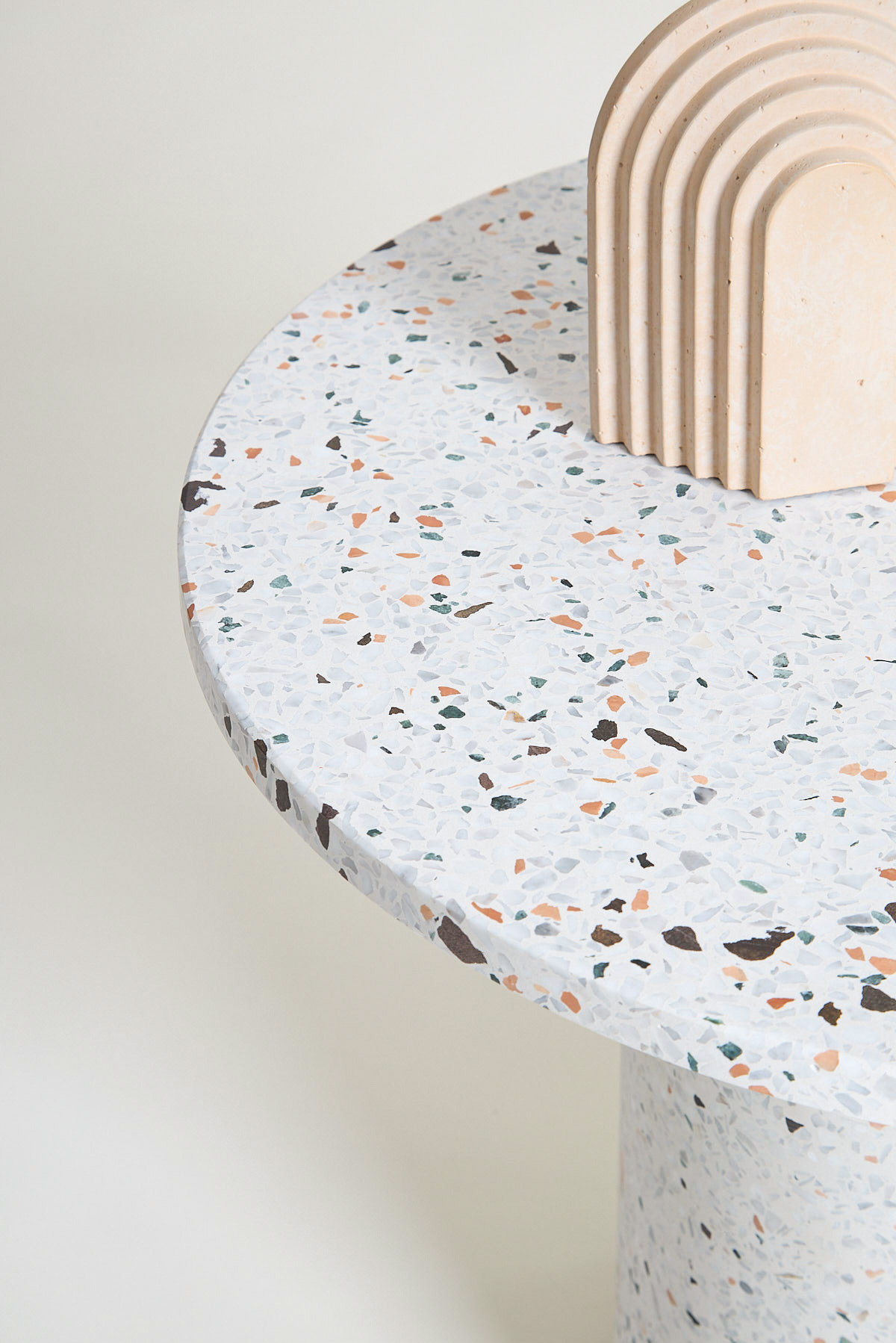 Fragment okrągłego stolika z blatem z lastryko w odcieniach bieli, szarości, brązu i pomarańczy, obok którego stoi dekoracyjny element architektoniczny w kształcie łuków z beżowego kamienia.