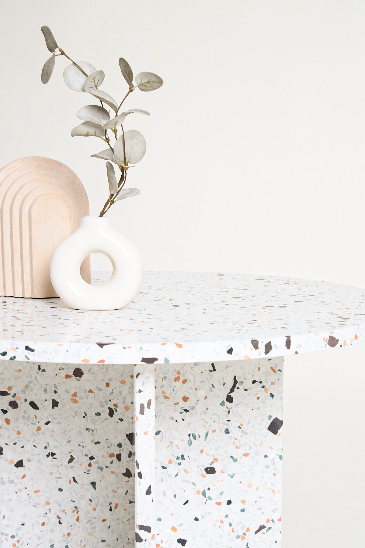 Stół z blatem i podstawą z lastryko, na nim ceramiczny wazon w kształcie pączka z gałązką eukaliptusa i dekoracyjna rzeźba w kształcie łuku.