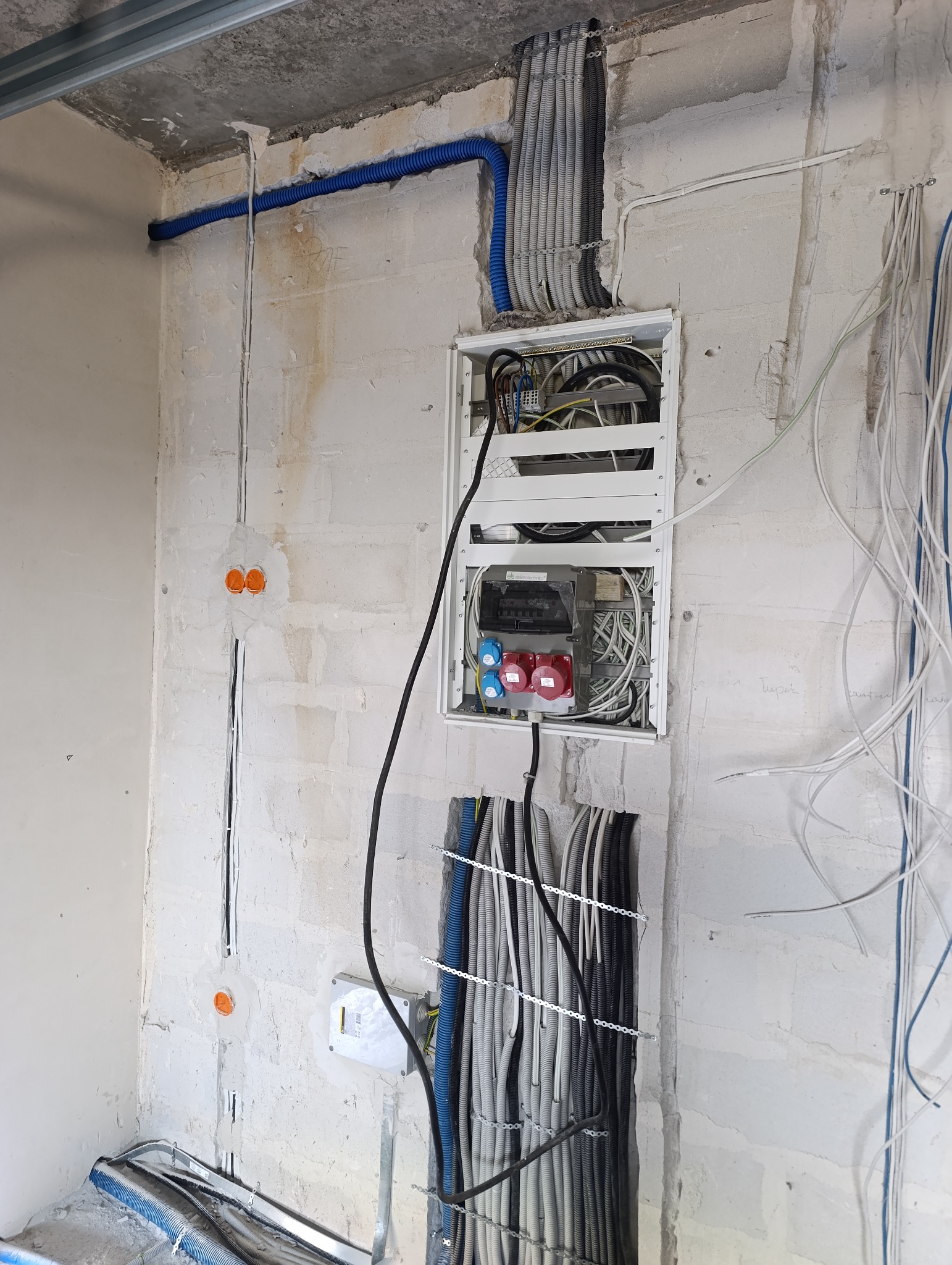 Rozdzielnica elektryczna z licznymi przewodami w ścianie z surowego betonu, widoczne peszle i kable w różnych kolorach, instalacja w trakcie montażu.