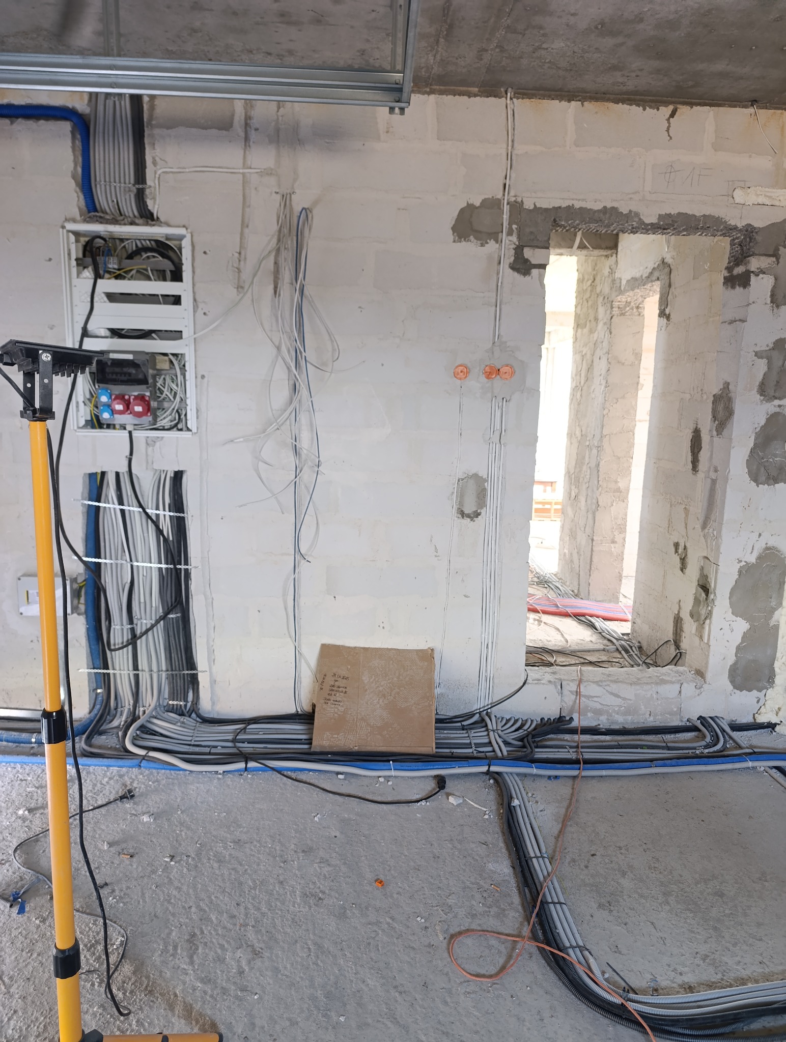 Rozprowadzenie instalacji elektrycznej w surowym wnętrzu. Widoczna skrzynka z bezpiecznikami, kable w peszlach i lampa budowlana. Prace w toku, stan deweloperski.