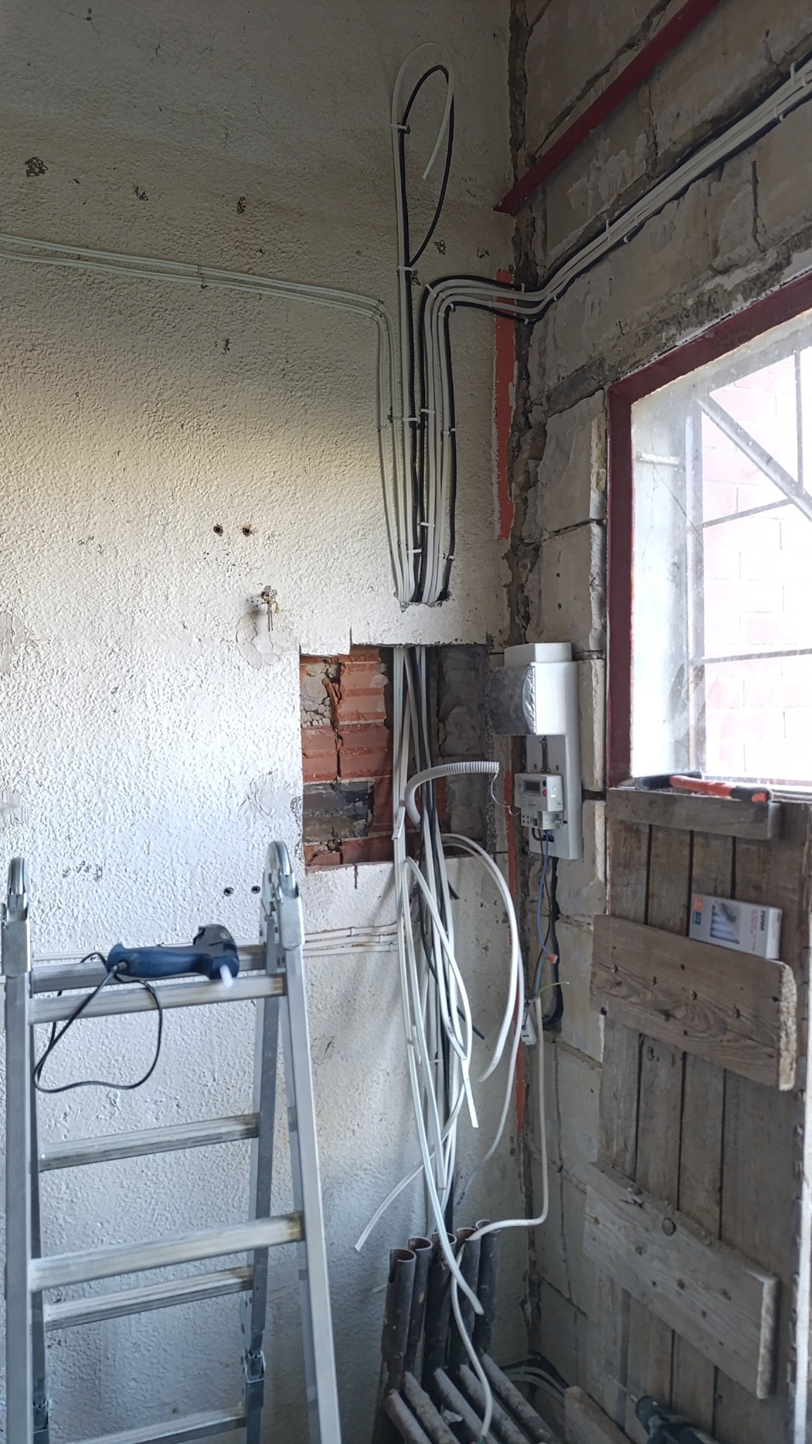 Wnętrze w trakcie prac elektrycznych: kable prowadzone po ścianach, częściowo w korytach, widoczna skrzynka elektryczna, drabina i narzędzia. Surowy stan wykończenia.