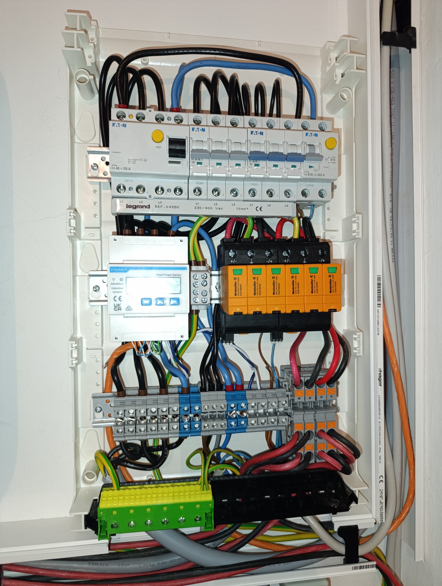 Rozdzielnica elektryczna w białej obudowie, widoczne zabezpieczenia Eaton, Legrand, Weidmüller, przewody w różnych kolorach. Elementy instalacji elektrycznej wewnątrz.