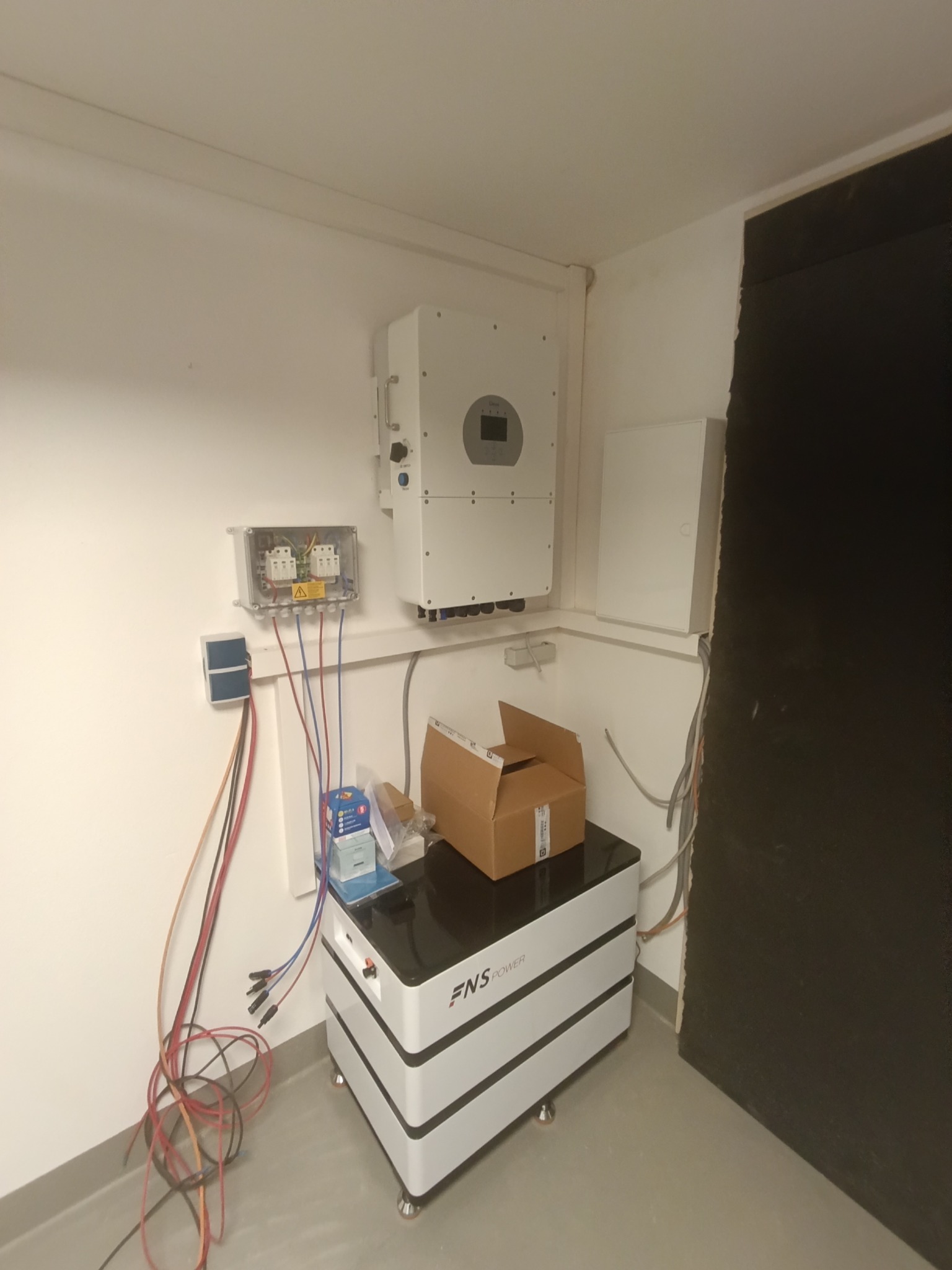 Biała bateria magazynu energii z podłączonymi przewodami, skrzynka z bezpiecznikami i falownik na ścianie w pomieszczeniu. Widoczne kable i akcesoria montażowe.