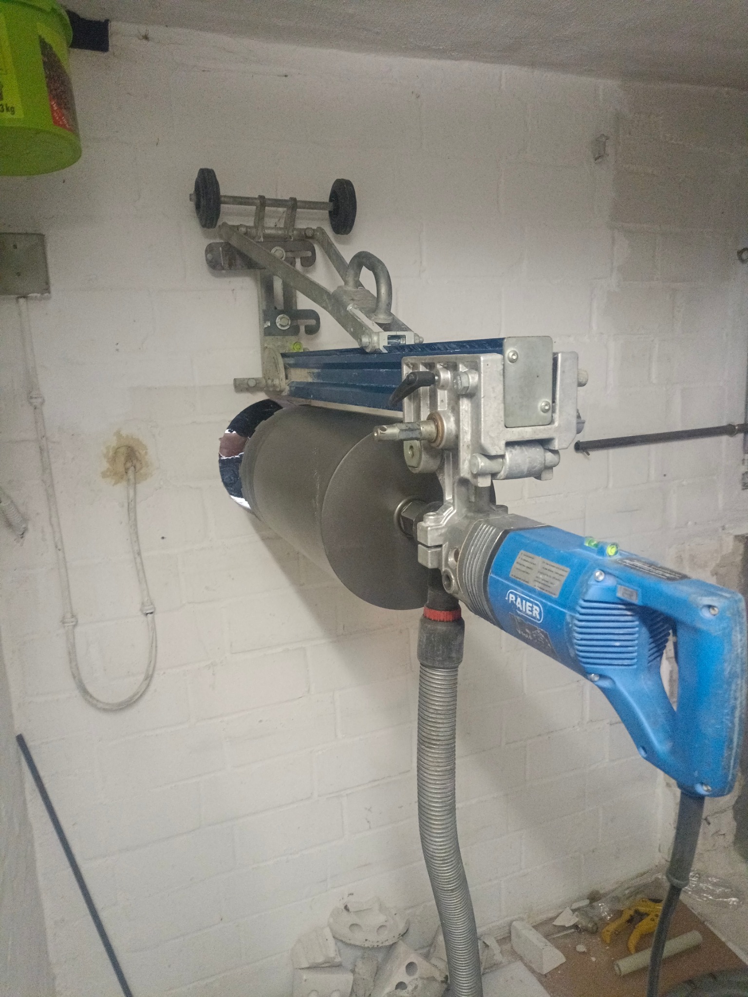 Wiertnica diamentowa marki Raier zamocowana do białej ściany z cegieł, przygotowująca otwór na rurę, z widocznymi śladami po poprzednich instalacjach i resztkami gruzu na podłodze.
