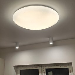 Szuba Volt System - Biały, okrągły plafon sufitowy z efektem rozproszonego światła, uzupełniony dwoma punktowymi lampami. Okno z białymi ramami w tle.