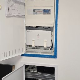 Szuba Volt System - Rozdzielnia elektryczna z otwartymi drzwiczkami, widoczne zabezpieczenia, licznik energii, router i multiswitch. Instalacja wewnątrz ściany, schludne okablowanie.