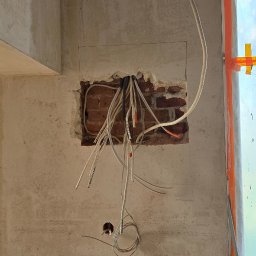 Szuba Volt System - Ściana z odsłoniętą cegłą i wystającymi przewodami elektrycznymi, widoczne narzędzia i drabina. Etap prac instalacyjnych w surowym wnętrzu.
