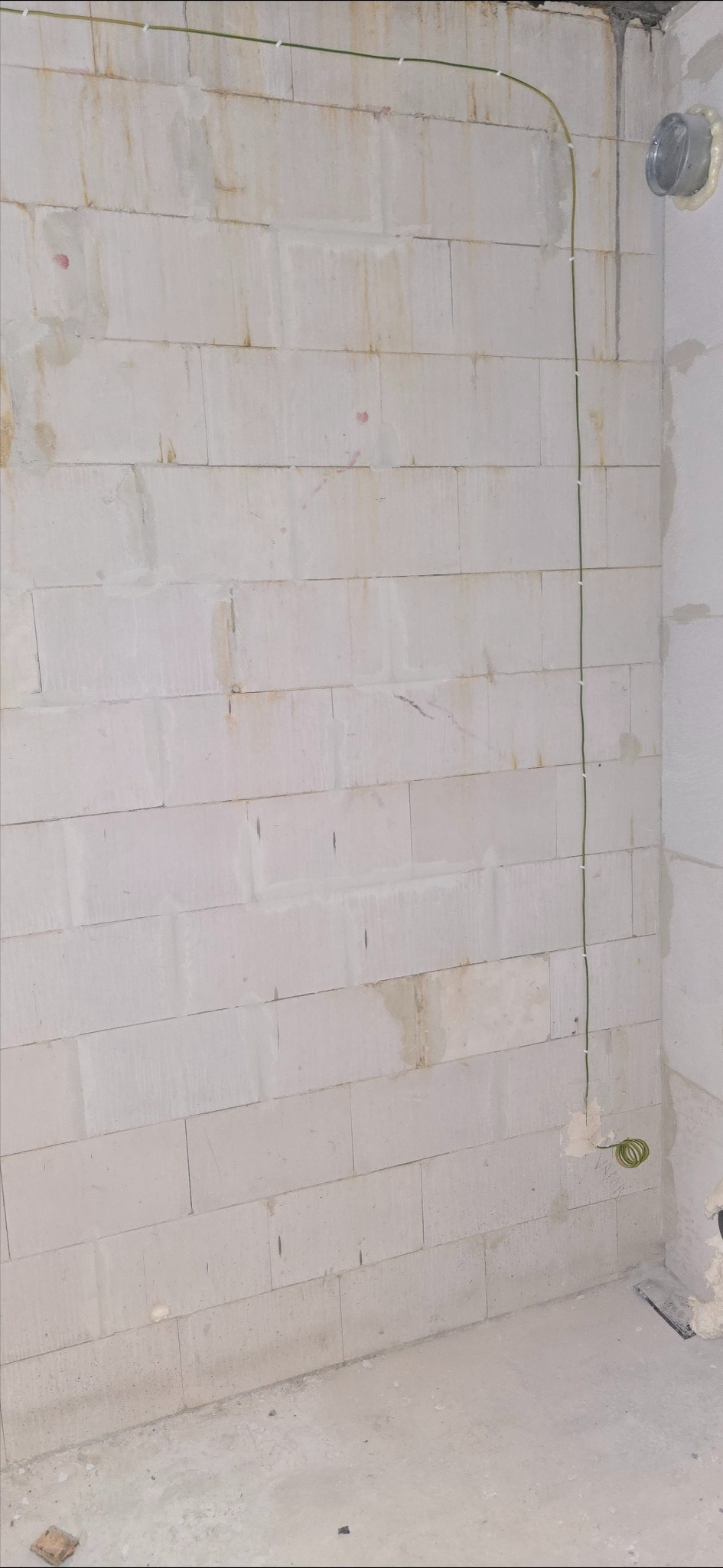 Ściana z betonu komórkowego z pionowo poprowadzoną, zieloną instalacją elektryczną, przymocowaną uchwytami. Widoczny fragment wentylacji w rogu.