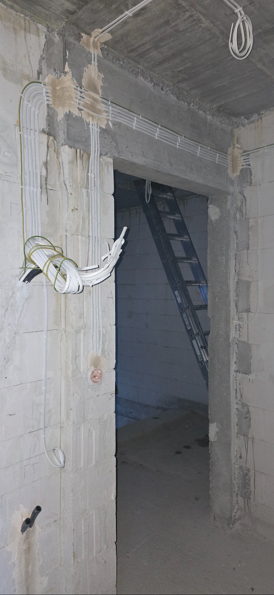 Instalacja elektryczna w surowym wnętrzu: białe kable prowadzone po ścianach z betonu komórkowego, widoczna drabina w przejściu. Etap prac przed wykończeniem.