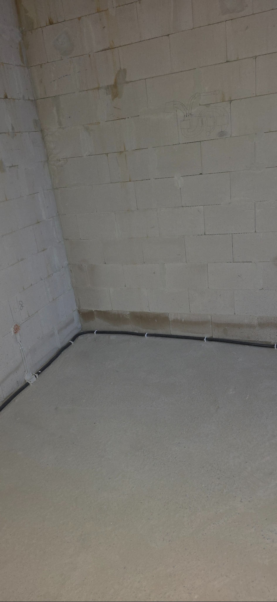 Szara, pusta przestrzeń z widoczną, czarną rurą instalacyjną przymocowaną do ściany z bloczków betonowych. Na ścianie widoczne szkice instalacji.