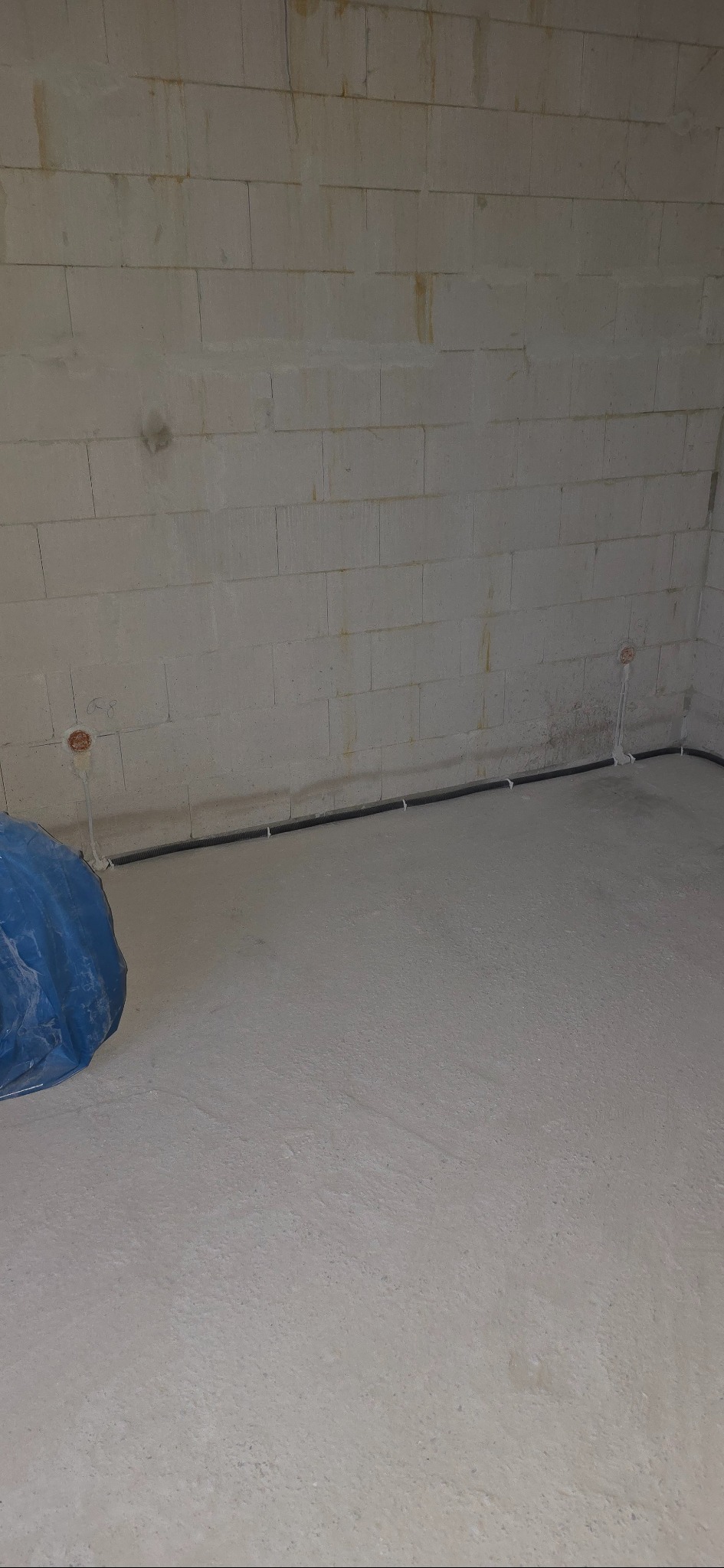 Surowy stan: czarna rura instalacji elektrycznej przy podłodze w jasnym, nieotynkowanym pomieszczeniu z bloków. Widoczne puszki instalacyjne.