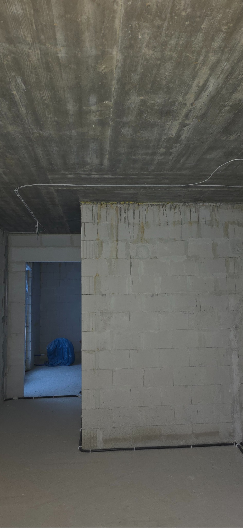 Surowy stan wnętrza w trakcie prac wykończeniowych. Widoczne przewody elektryczne poprowadzone po ścianach i suficie, surowy betonowy strop, ściany z bloczków.