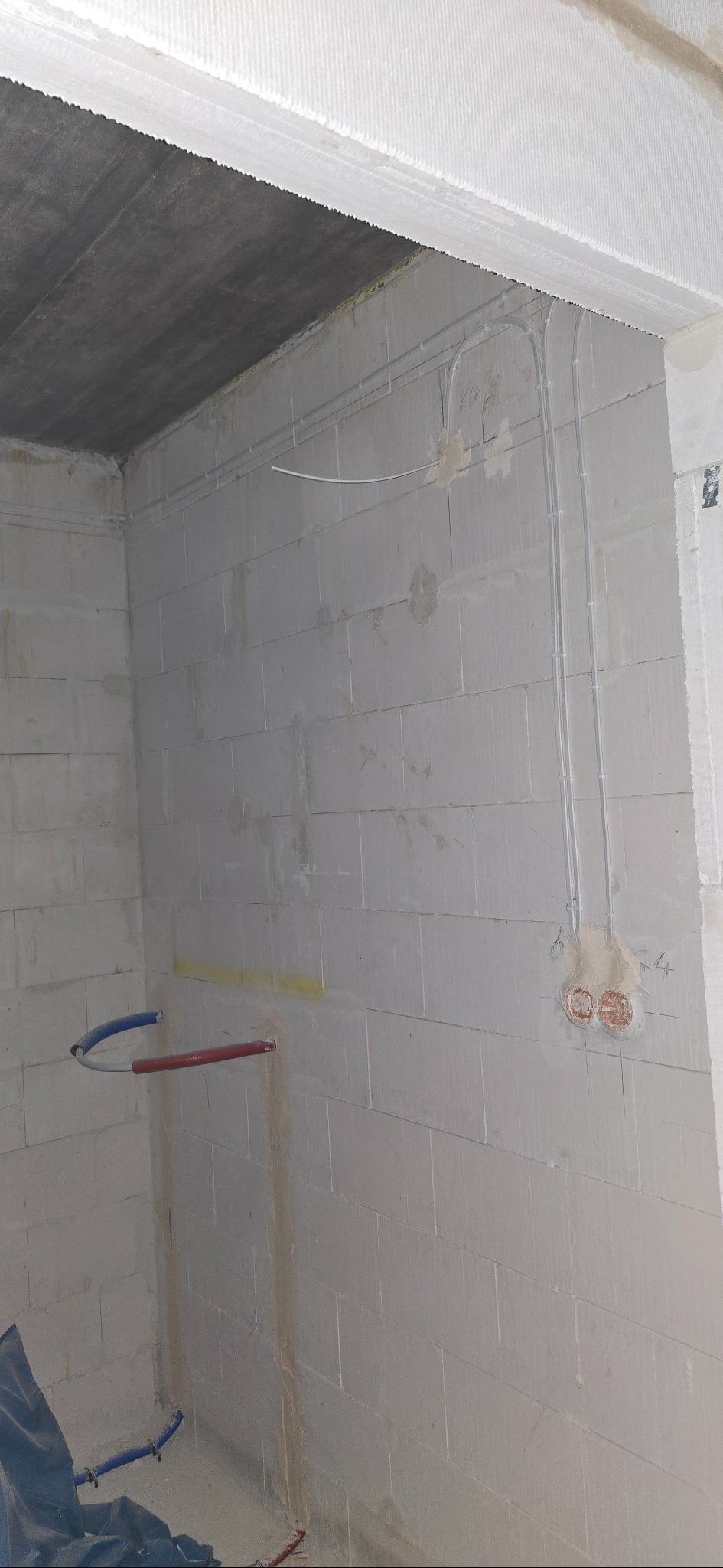 Surowa ściana z betonu komórkowego z widocznymi przewodami elektrycznymi w peszlach i puszkami instalacyjnymi. Fragment instalacji wodnej z rur PEX.
