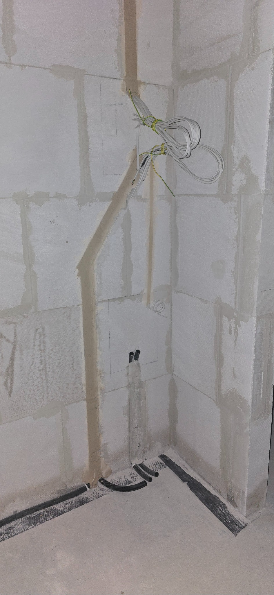 Ściana z instalacją elektryczną w trakcie montażu: przewody w peszlach ułożone w bruzdach, kable zwisają ze ściany. Widoczne puszki pod gniazdka. Surowy stan.