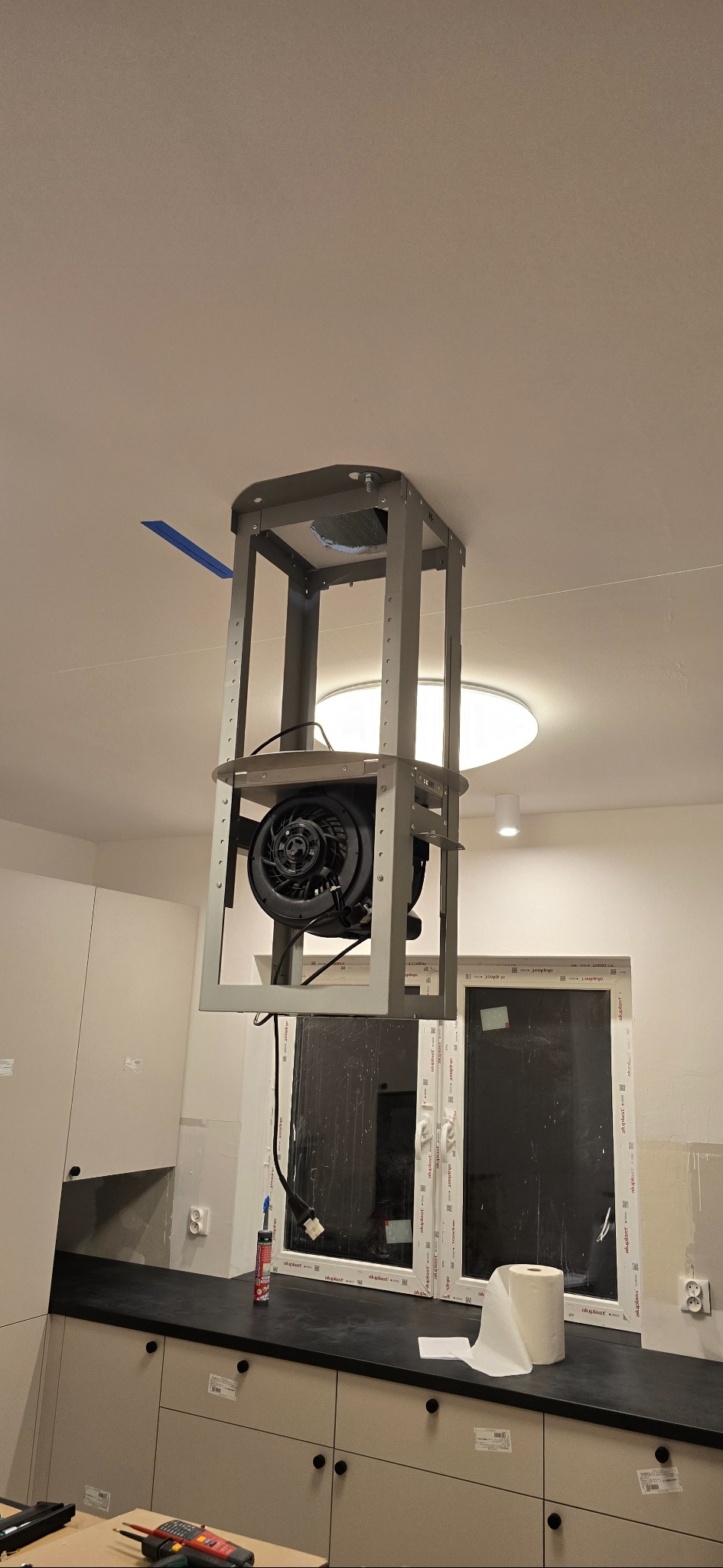 Montaż wentylatora sufitowego w kuchni, widoczny metalowy stelaż i czarny wentylator. W tle okno i zabudowa kuchenna. Etap instalacji, widoczne narzędzia.