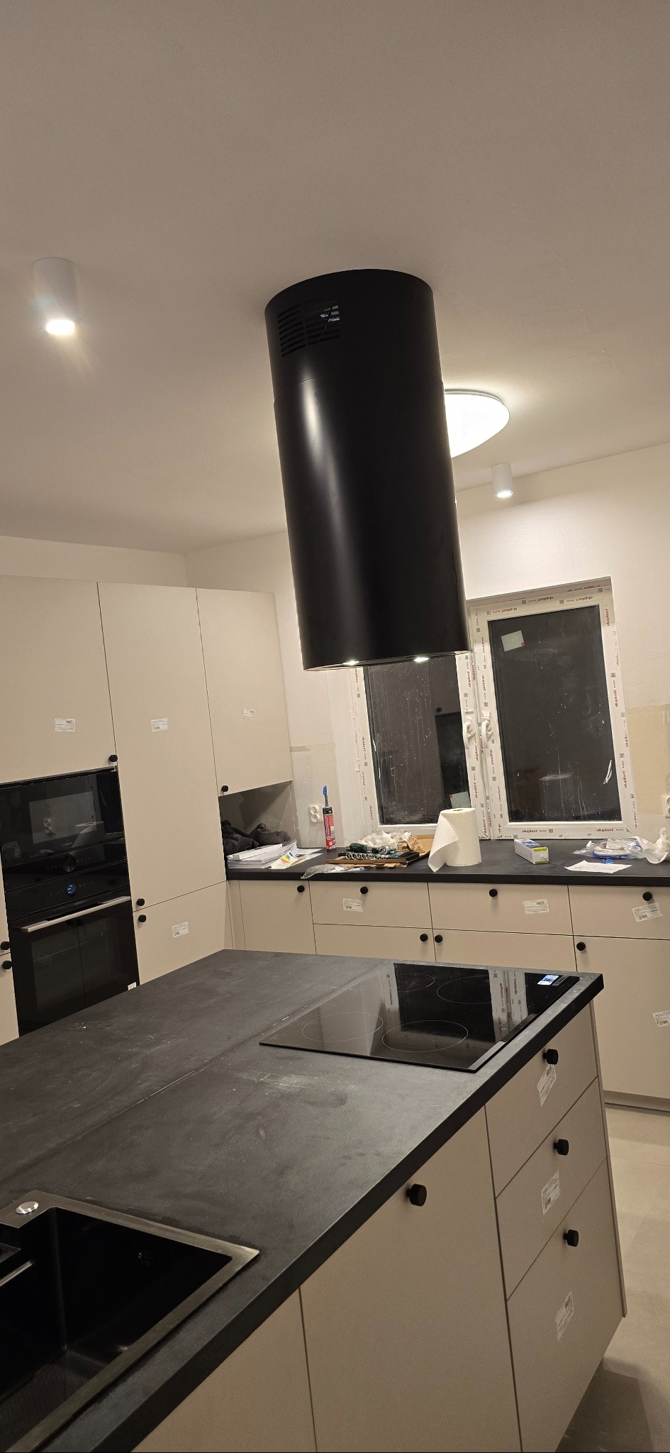 Nowoczesna kuchnia w trakcie montażu: czarny okrągły okap nad wyspą z płytą indukcyjną. Widoczne szafki i okno. Oświetlenie sufitowe.