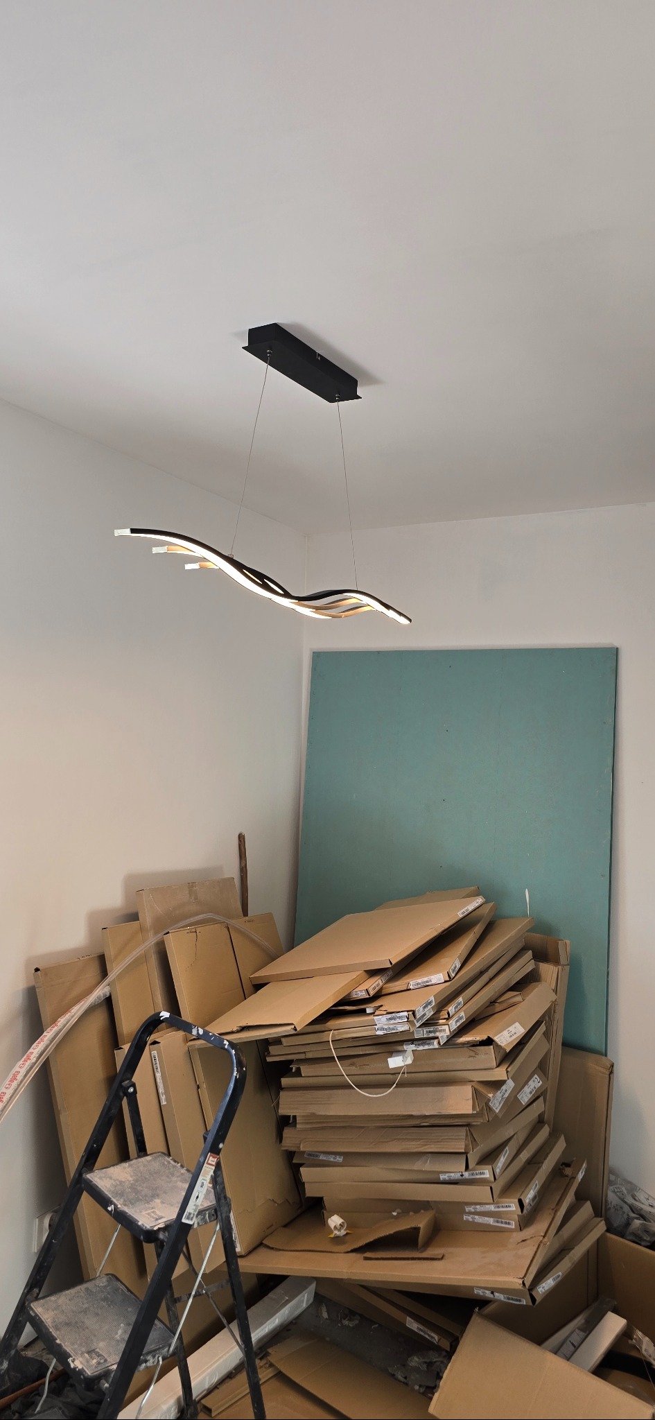 Nowoczesna, falista lampa LED zamontowana na suficie w pomieszczeniu w trakcie remontu. W tle sterta kartonów i drabina. Ujęcie z dołu, podkreślające design lampy.