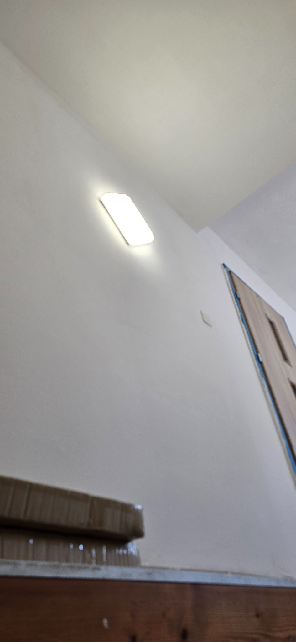 Nowoczesna, prostokątna lampa LED na białej ścianie w korytarzu, oświetlająca przestrzeń. W tle widoczne drzwi i fragment schodów.