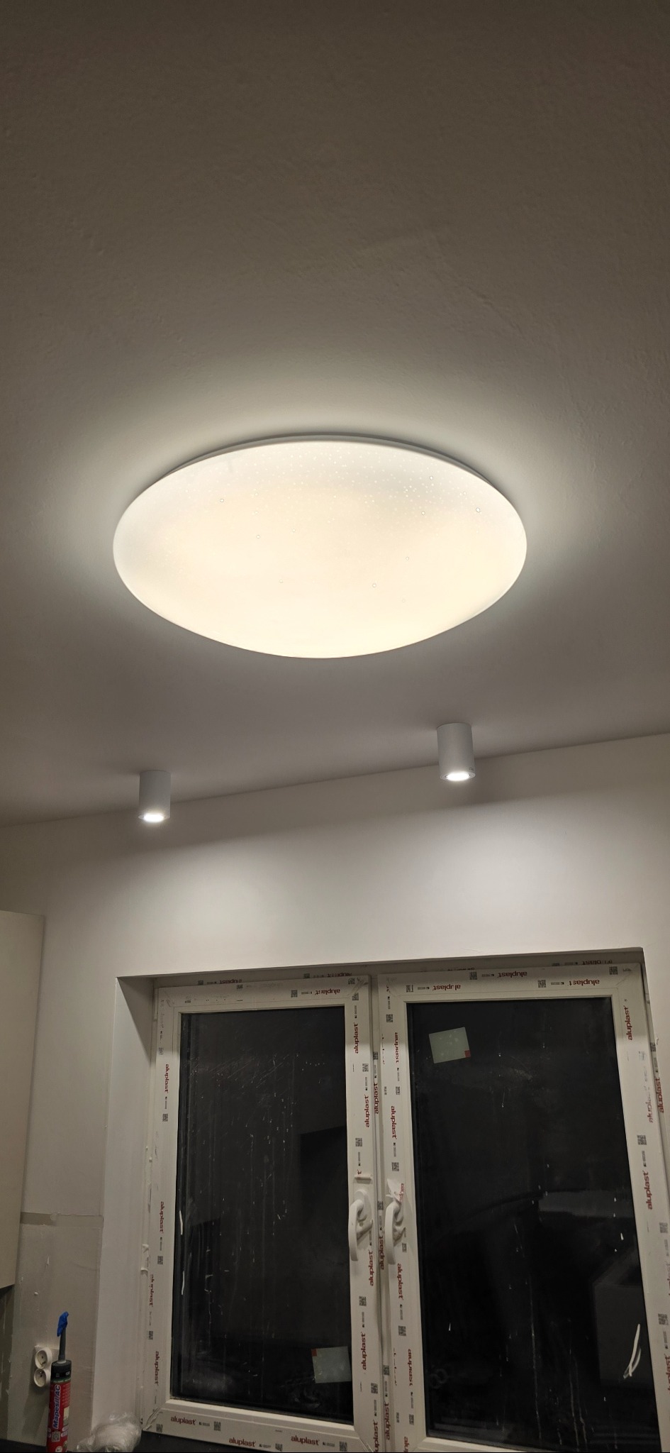 Biały, okrągły plafon sufitowy z efektem rozproszonego światła, uzupełniony dwoma punktowymi lampami. Okno z białymi ramami w tle.