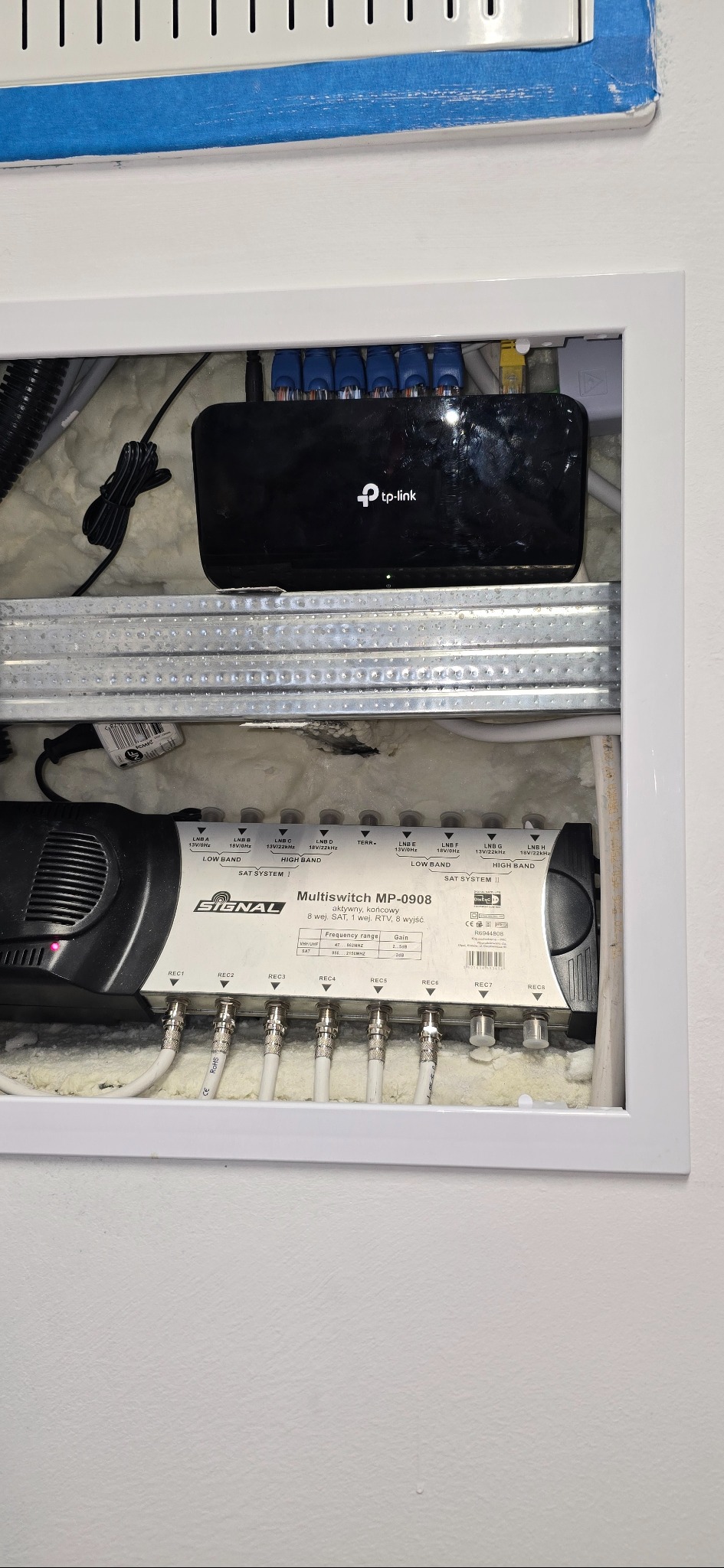 Rozdzielacz sygnału SAT Signal MP-0908 z podłączonymi kablami koncentrycznymi, router TP-Link i kable sieciowe w białej wnęce. Widoczne złącza i oznaczenia.
