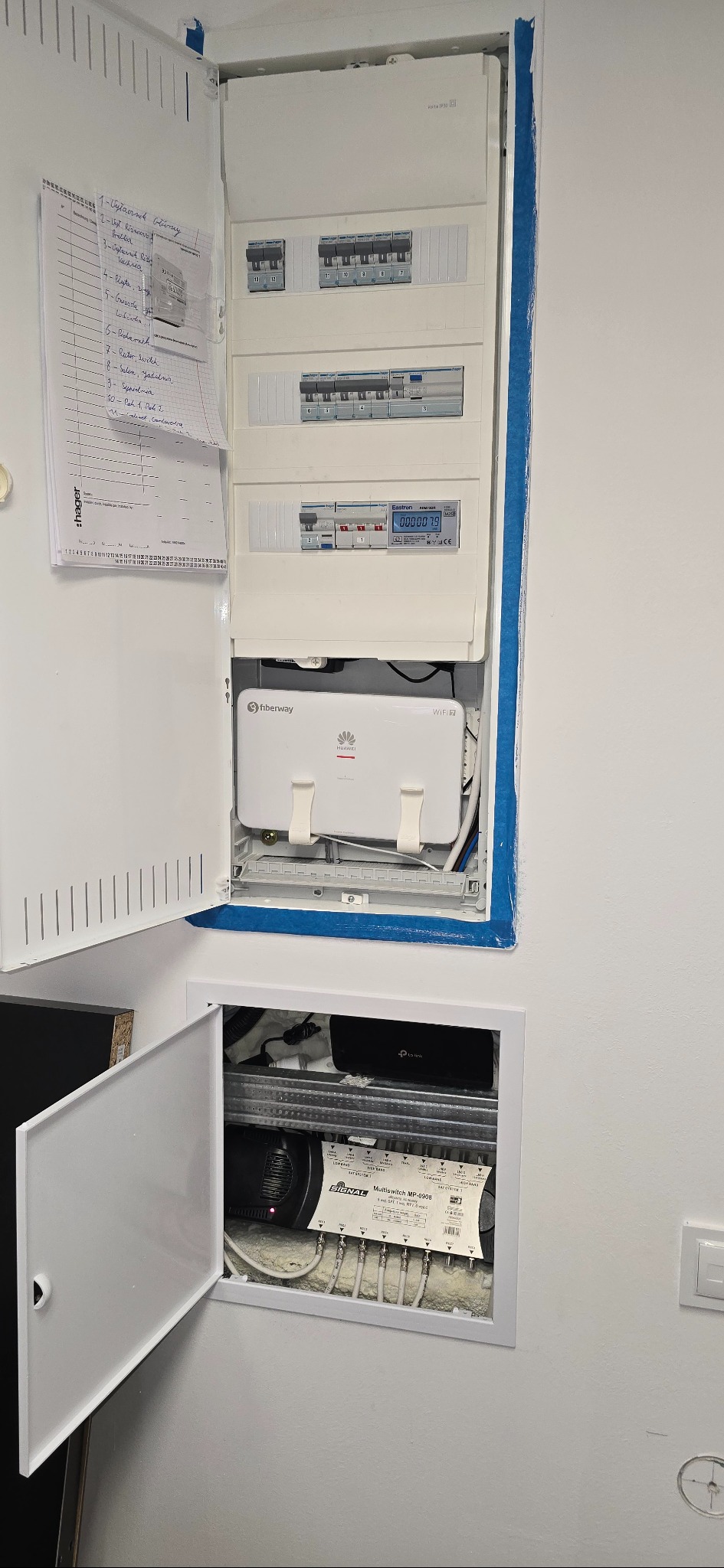 Rozdzielnia elektryczna z otwartymi drzwiczkami, widoczne zabezpieczenia, licznik energii, router i multiswitch. Instalacja wewnątrz ściany, schludne okablowanie.