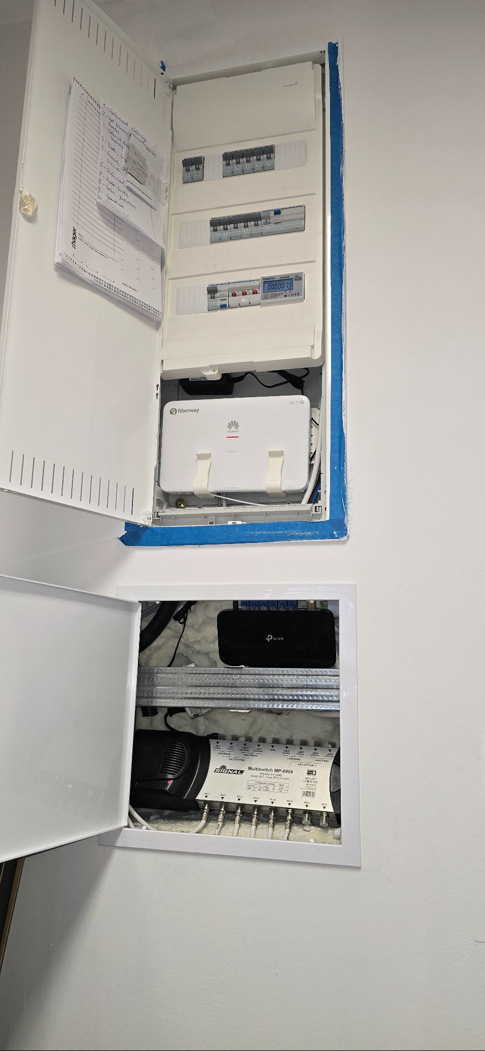 Otwarta skrzynka rozdzielcza z bezpiecznikami, routerem Fiberway, TP-Link i multiswitchem Signal MP-0908. Widoczne kable i lista kontrolna na drzwiach.