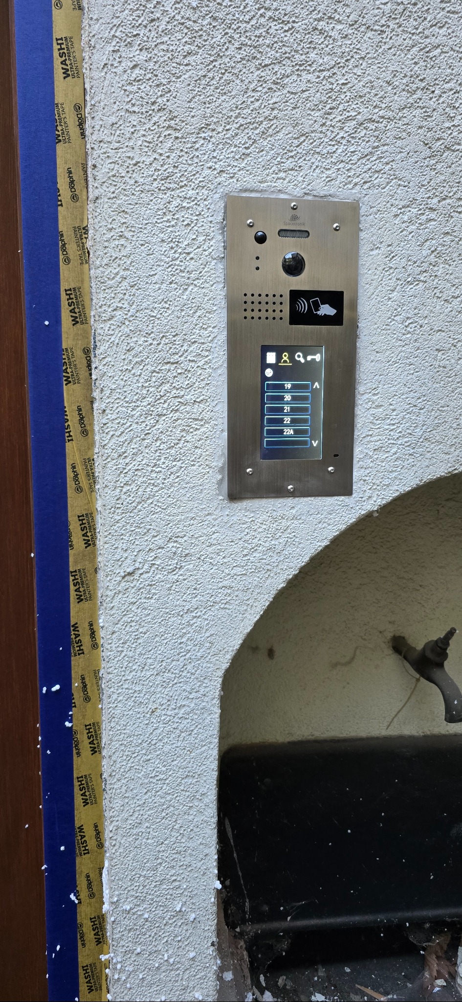Panel domofonowy z ekranem LCD, zamontowany na teksturowanej ścianie zewnętrznej. Widoczne numery mieszkań i ikony funkcji. Taśma malarska zabezpiecza krawędź ściany.