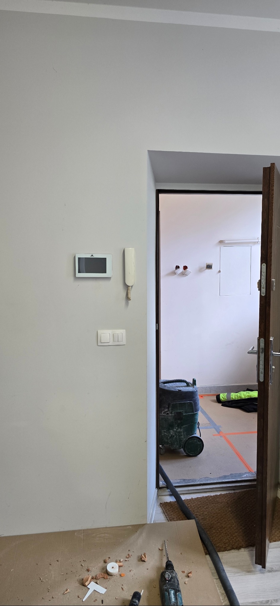 Wnętrze mieszkania w trakcie prac wykończeniowych. Widoczny interkom, włączniki światła, odkurzacz przemysłowy i narzędzia na podłodze zabezpieczonej kartonem.