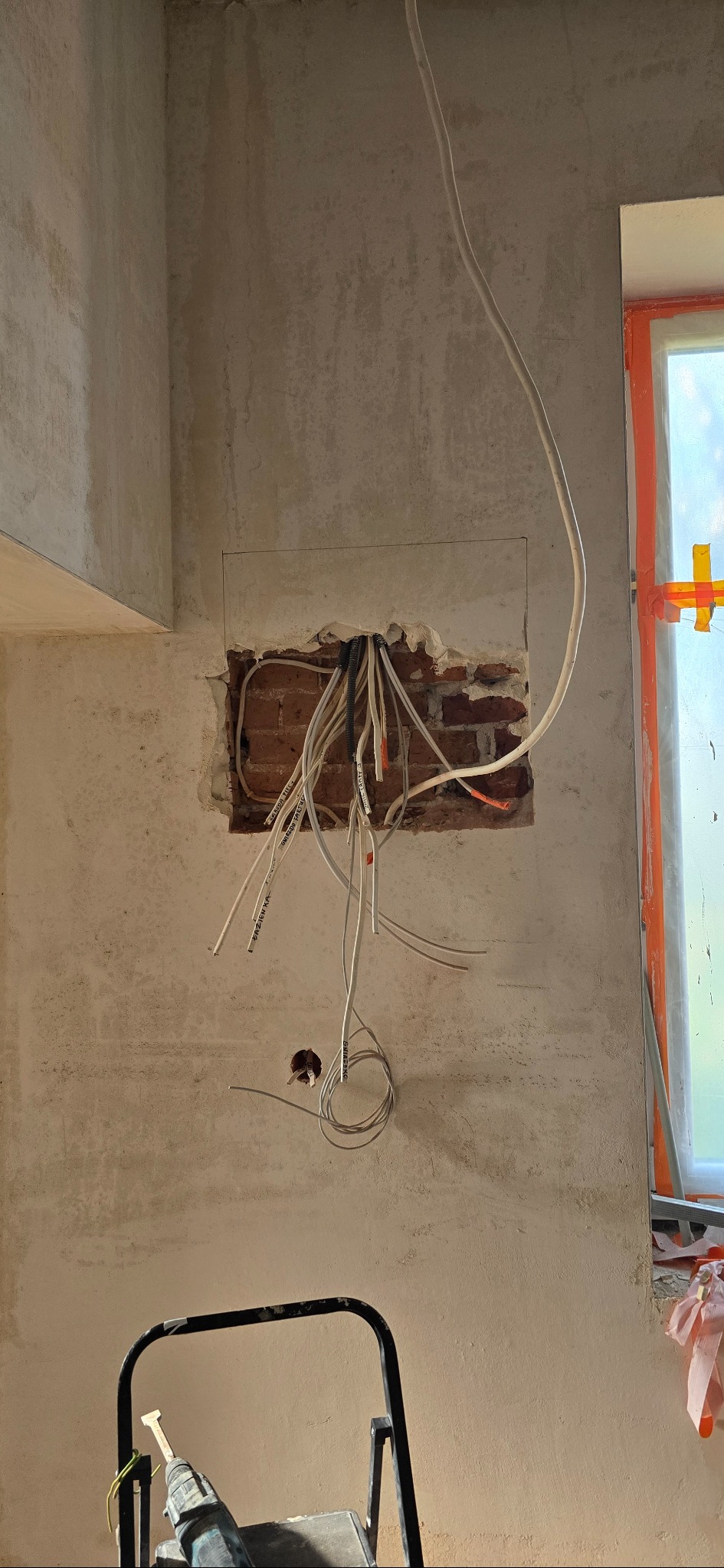Ściana z odsłoniętą cegłą i wystającymi przewodami elektrycznymi, widoczne narzędzia i drabina. Etap prac instalacyjnych w surowym wnętrzu.