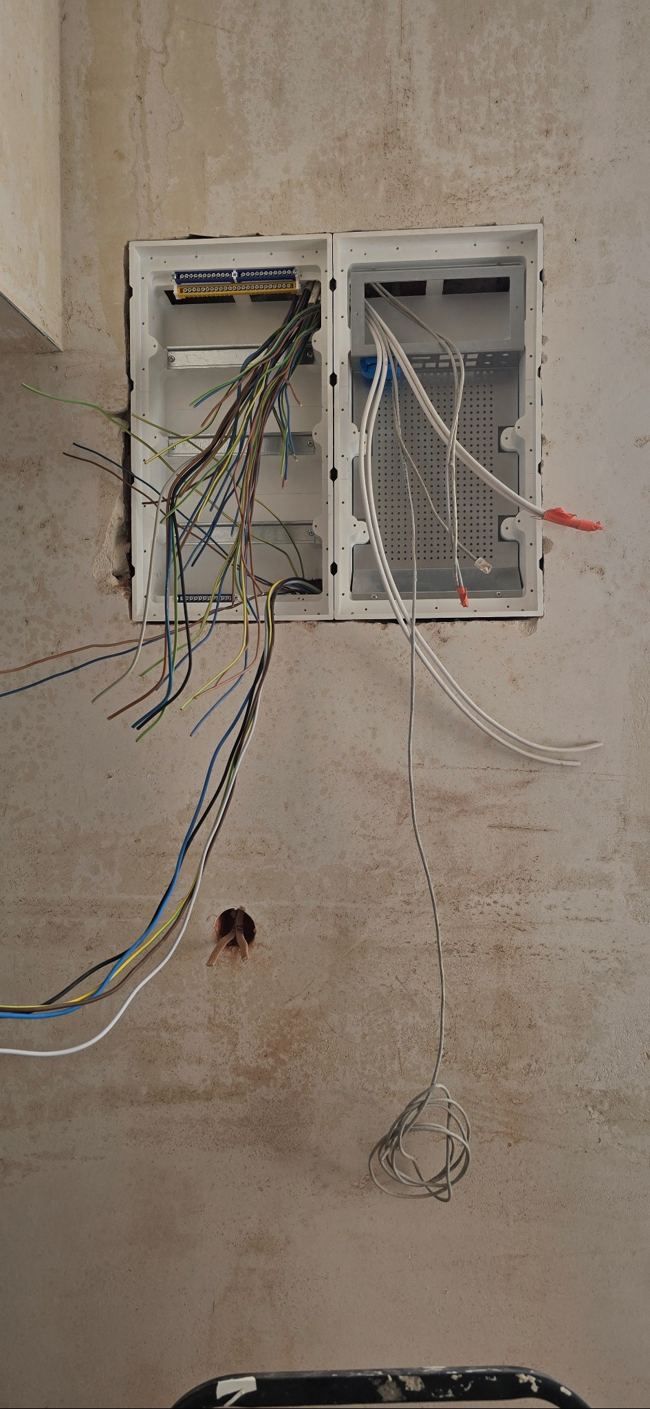 Dwa białe panele elektryczne w ścianie z wystającymi przewodami w różnych kolorach. Jeden panel z listwą zaciskową. Surowa ściana w tle.