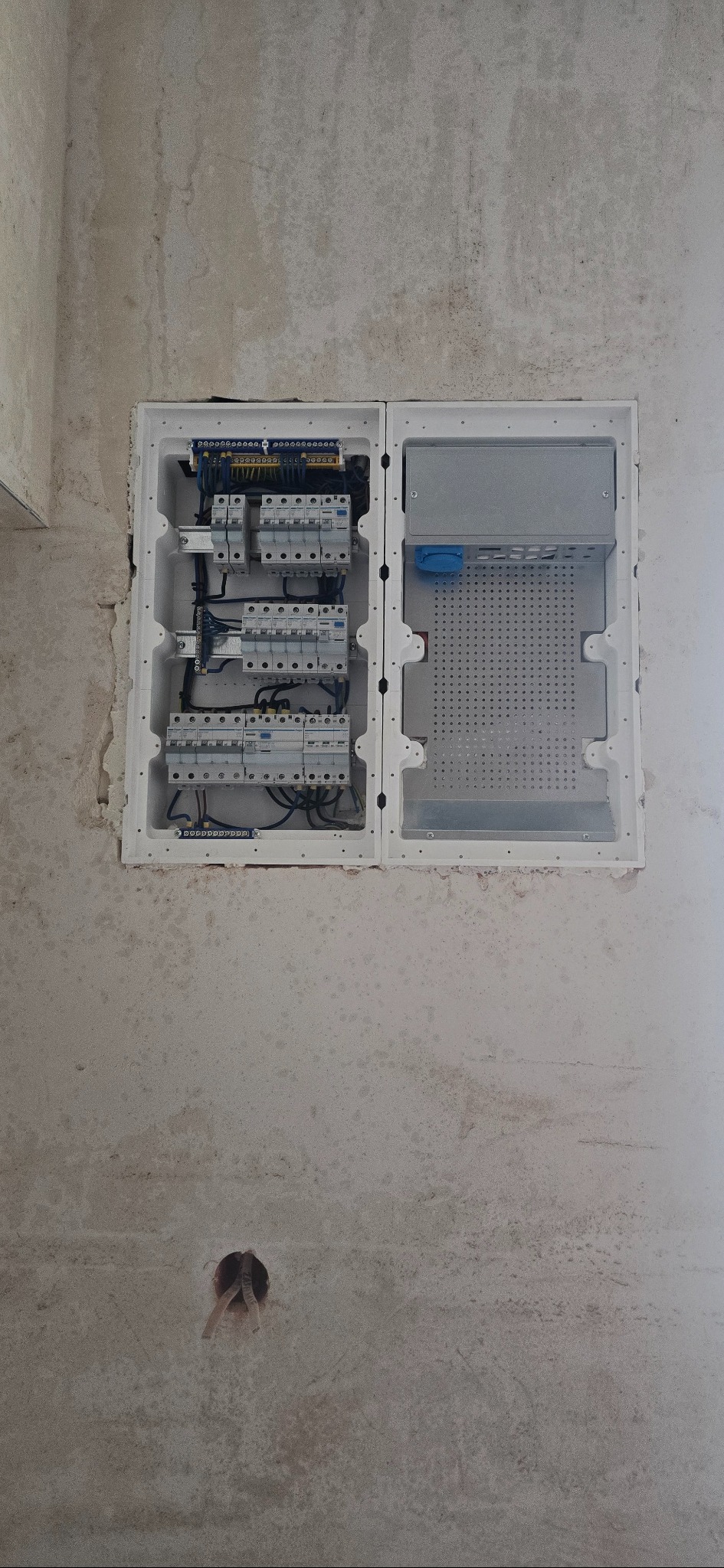 Otwarta, biała skrzynka rozdzielcza z bezpiecznikami i okablowaniem, zamontowana na surowej, szarej ścianie w trakcie prac wykończeniowych. Widoczny otwór w ścianie poniżej.