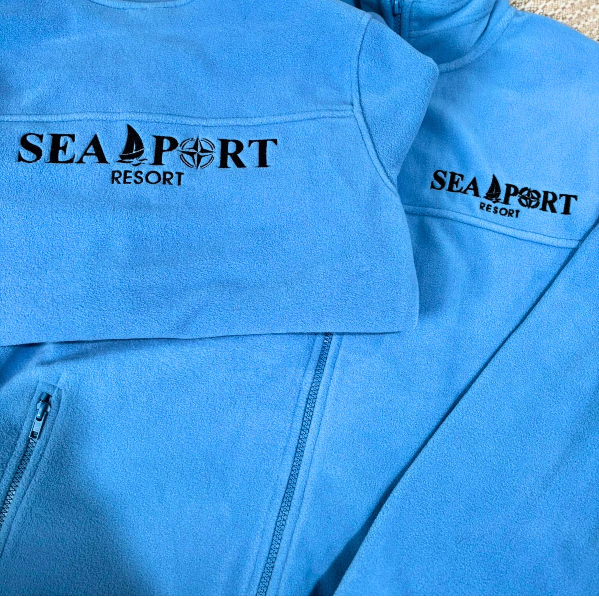 Dwie błękitne bluzy polarowe z haftowanym logo 'SEA PORT RESORT', prezentujące precyzję i jakość wykonania haftu.