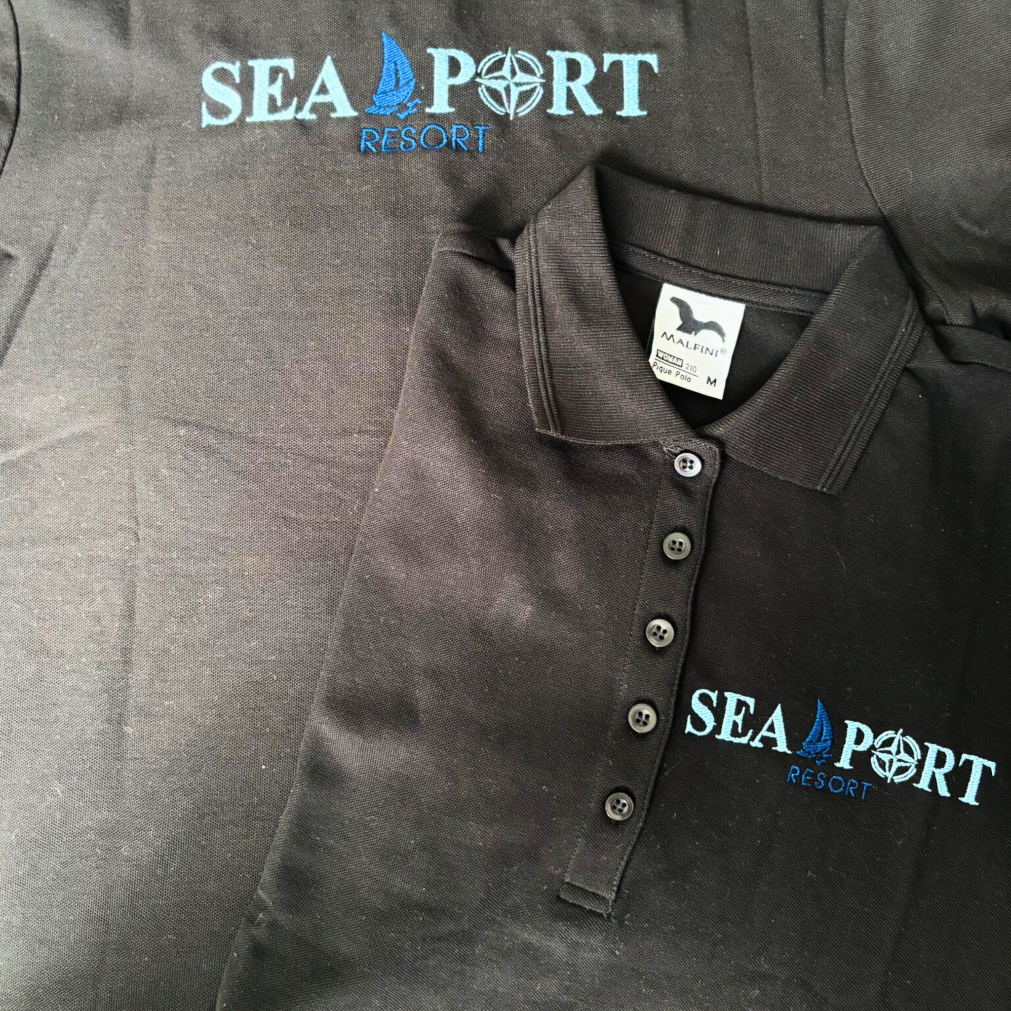 Dwie ciemne koszulki polo z błękitnym, maszynowym wzorem 'SEA PORT RESORT', jedna z metką. Widoczny detal wzoru z motywem żaglówki i kompasu.