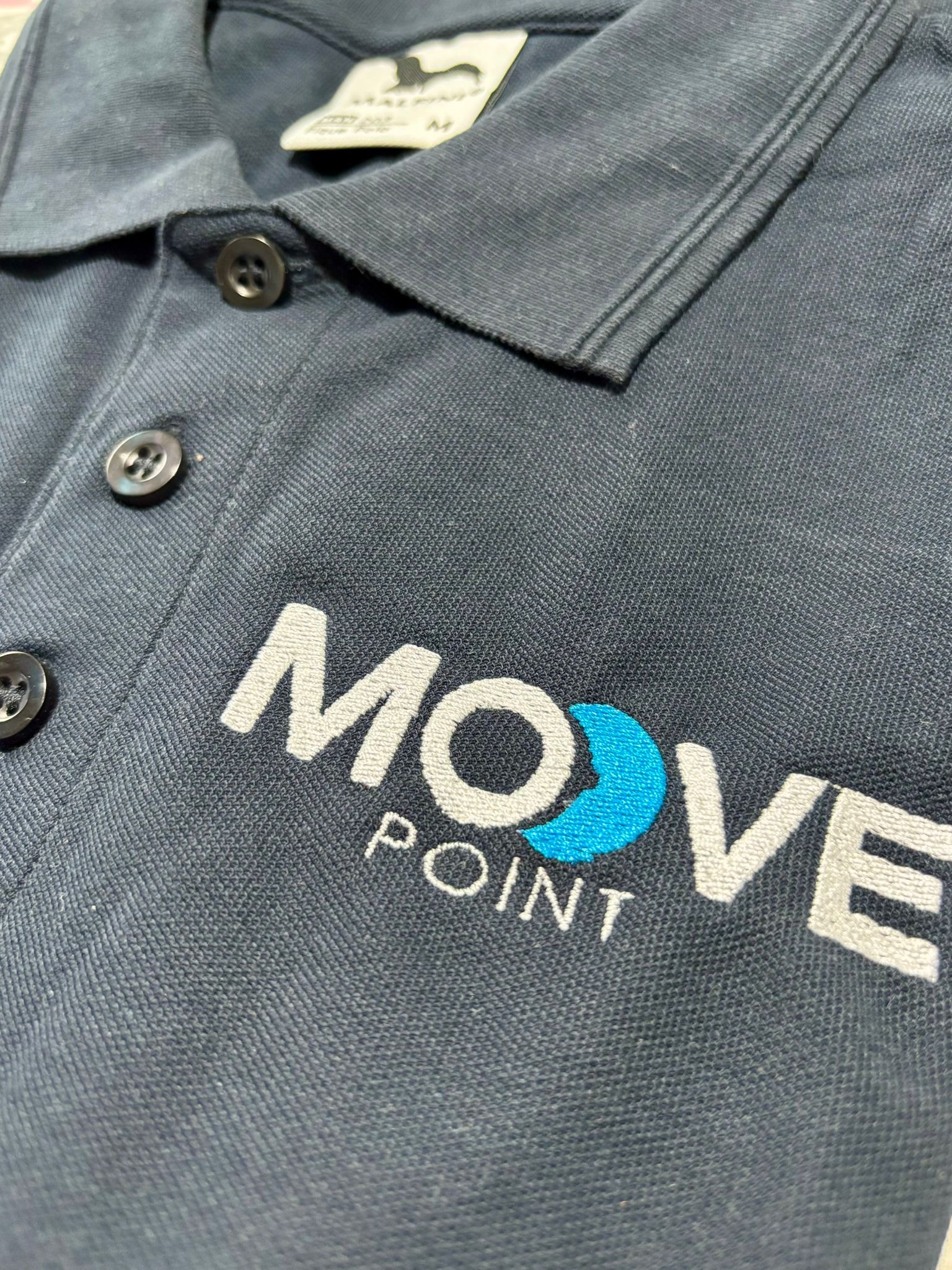 Granatowa koszulka polo z wyhaftowanym logo 'MOVE POINT' w kolorze białym i turkusowym. Widoczne guziki i metka marki. Detal haftu, zbliżenie na logo.