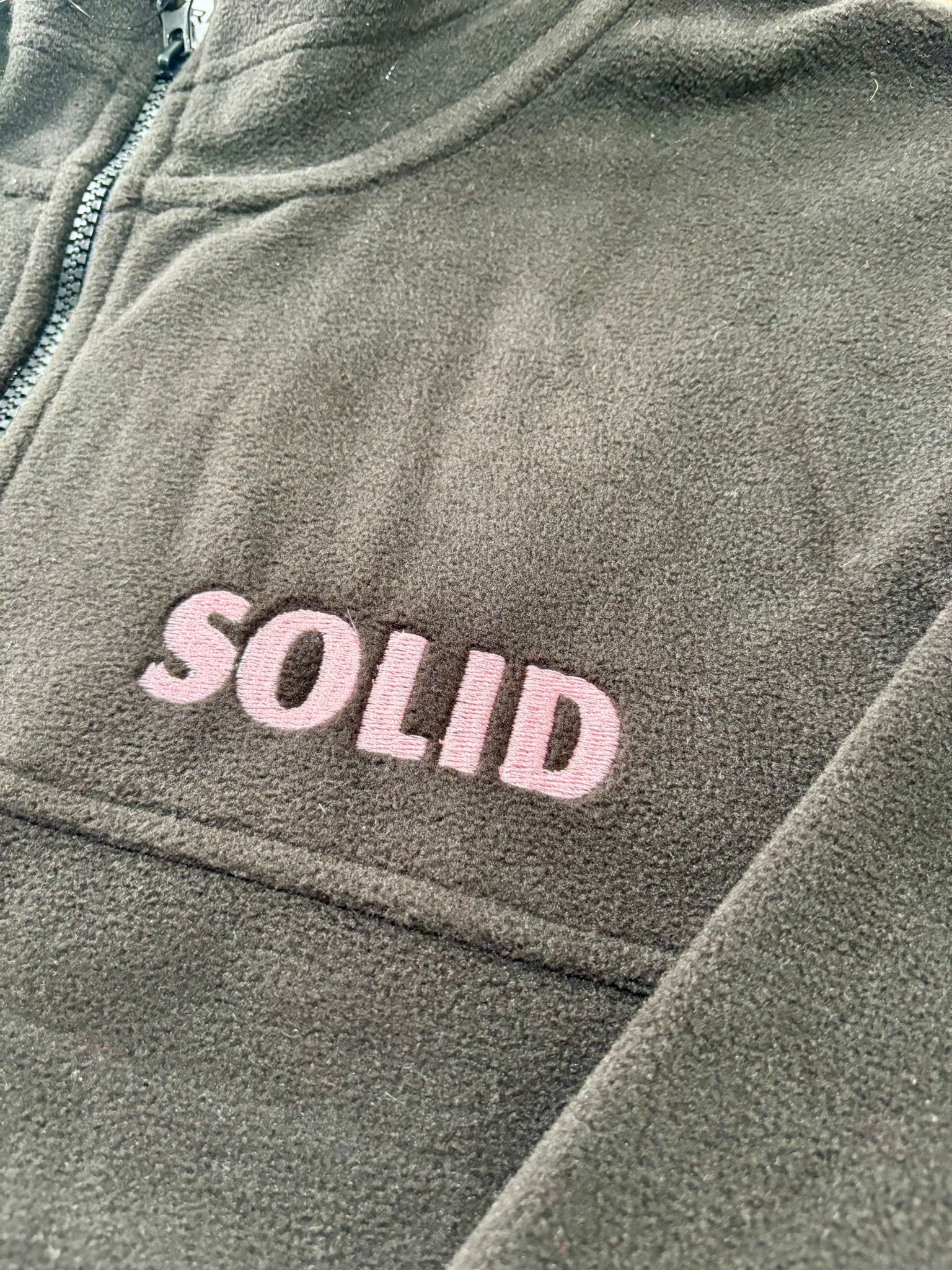 Ciemnoszara bluza polarowa z różowym napisem 'SOLID' wykonanym techniką haftu. Widoczny zamek błyskawiczny i faktura materiału.