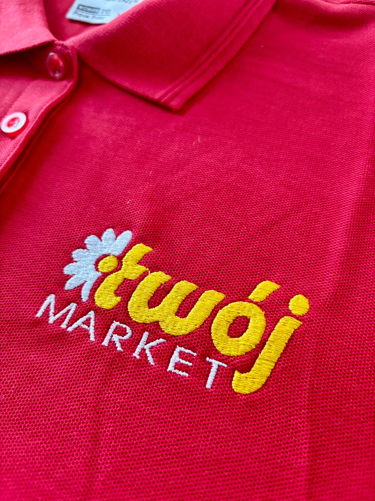 Czerwona koszulka polo z haftowanym logo 'Twój Market' w kolorze żółto-białym. Detalowy widok na haft, ukazujący precyzję wykonania na materiale koszulki.