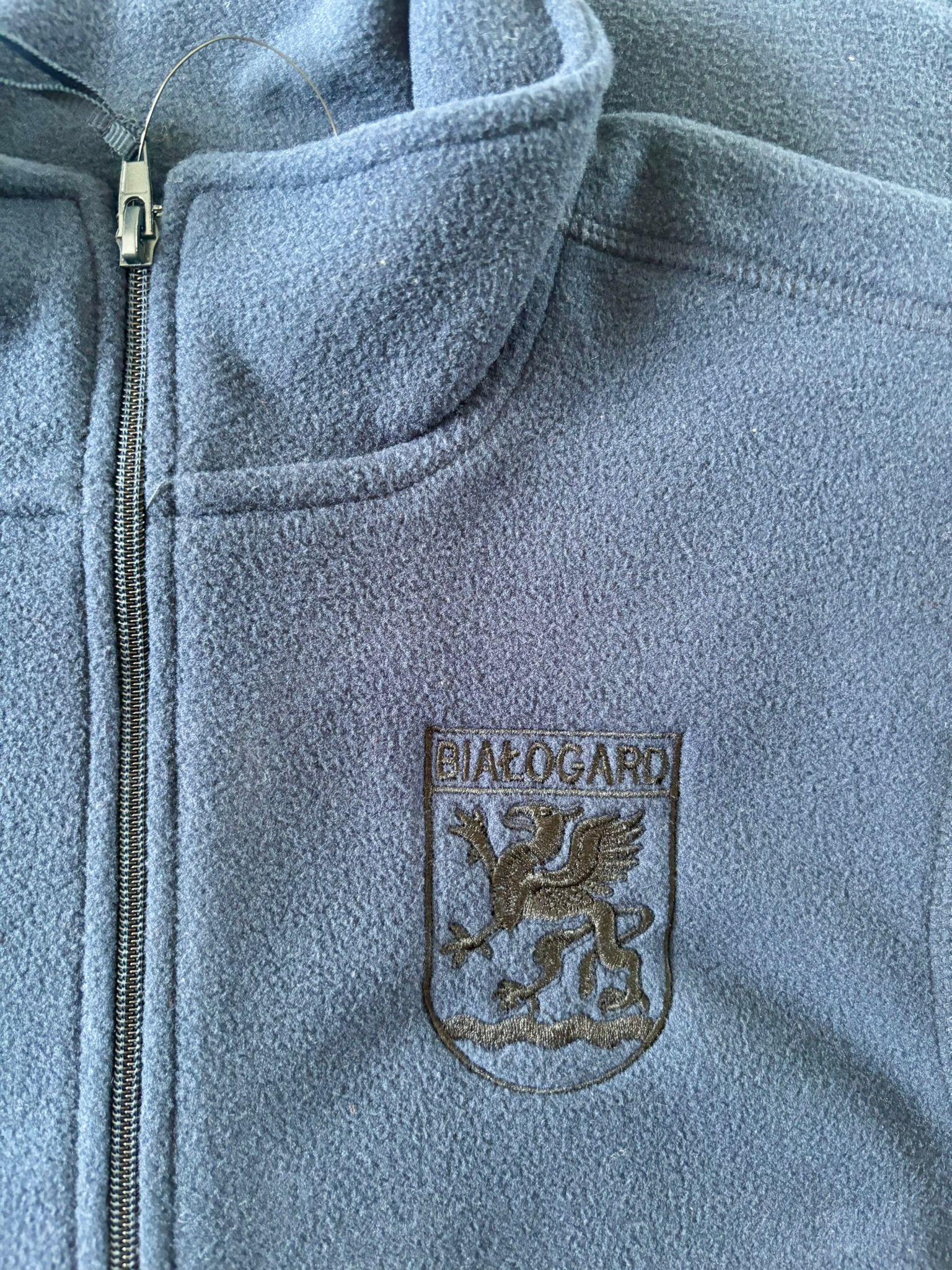 Błękitna bluza polarowa z zamkiem i wyhaftowanym herbem Białogardu, ukazująca detal precyzyjnego wykonania i wysoką jakość materiału.